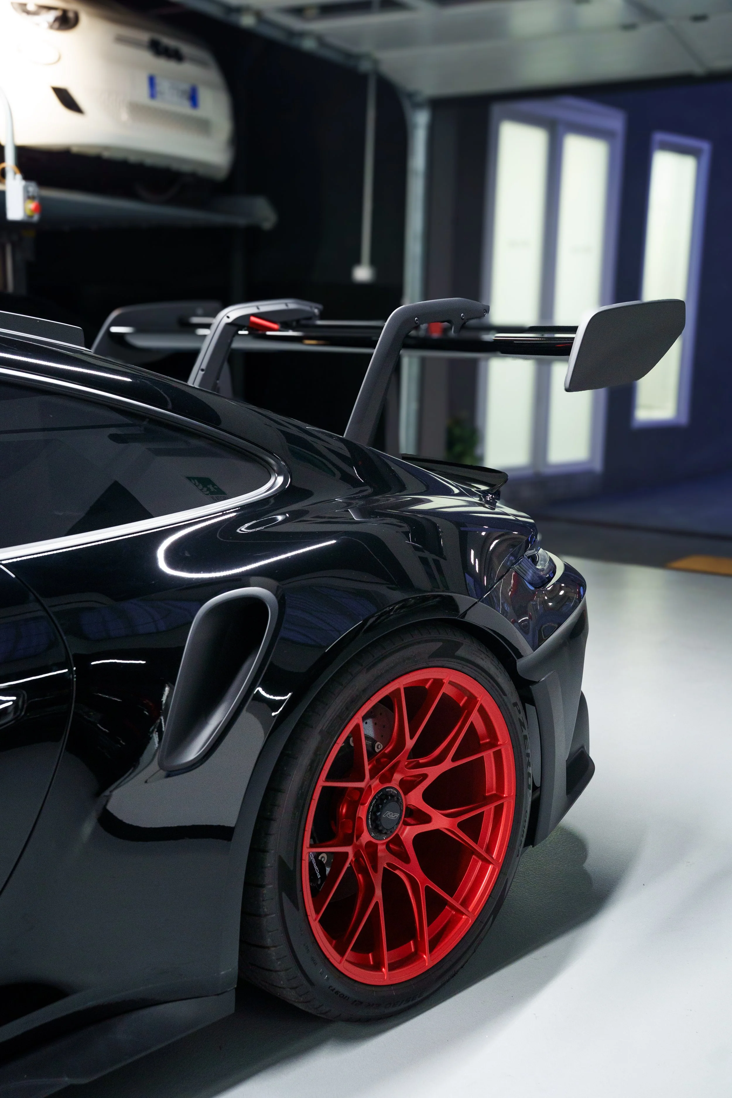 Close-up of a black sports car with red wheels and a rear wing, parked in a garage with a white car on a lift in the background.