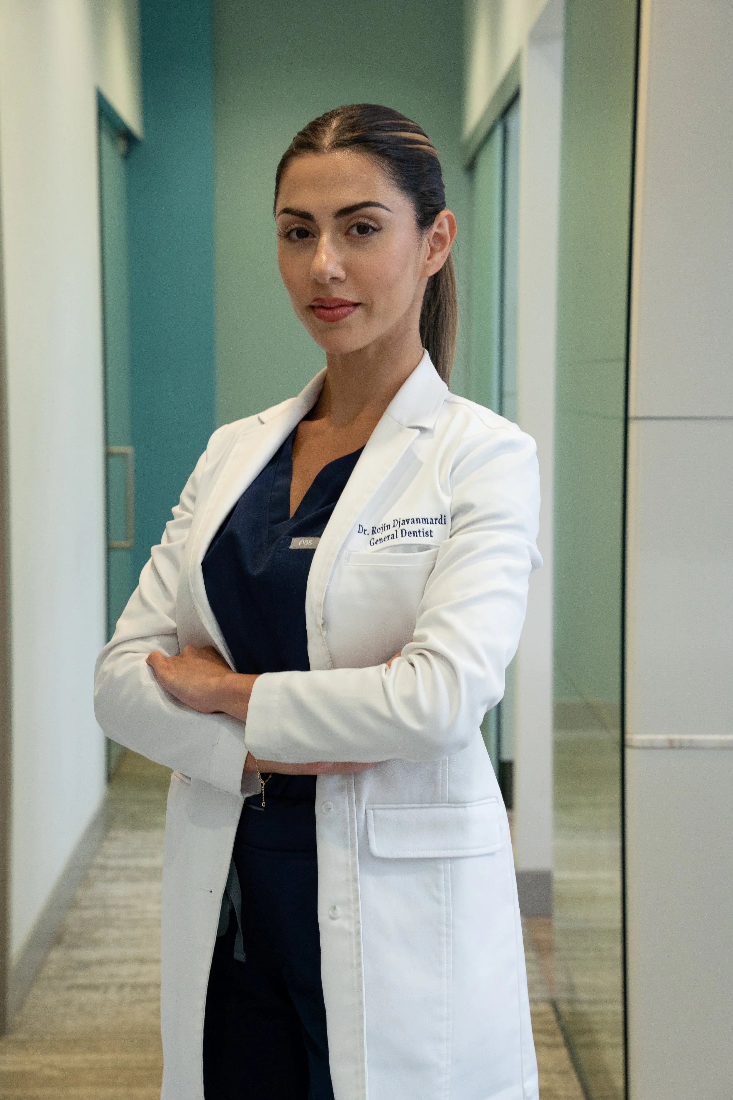 A female doctor stands in a hospital corridor with crossed arms, wearing a white coat over navy scrubs, with a confident expression.