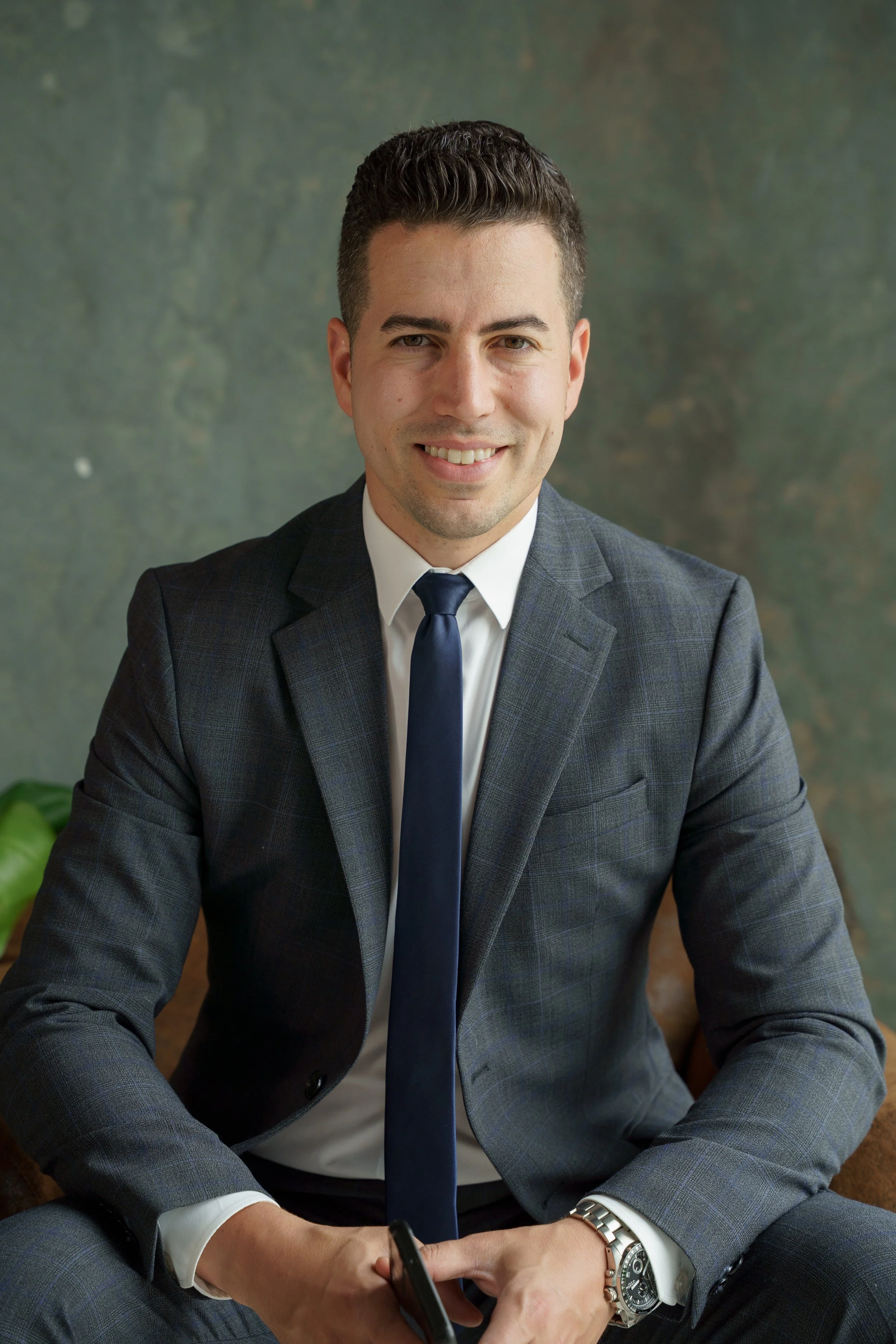 A man in a dark gray suit and white shirt, wearing a dark blue tie, sitting on a brown chair with a green plant visible in the background.