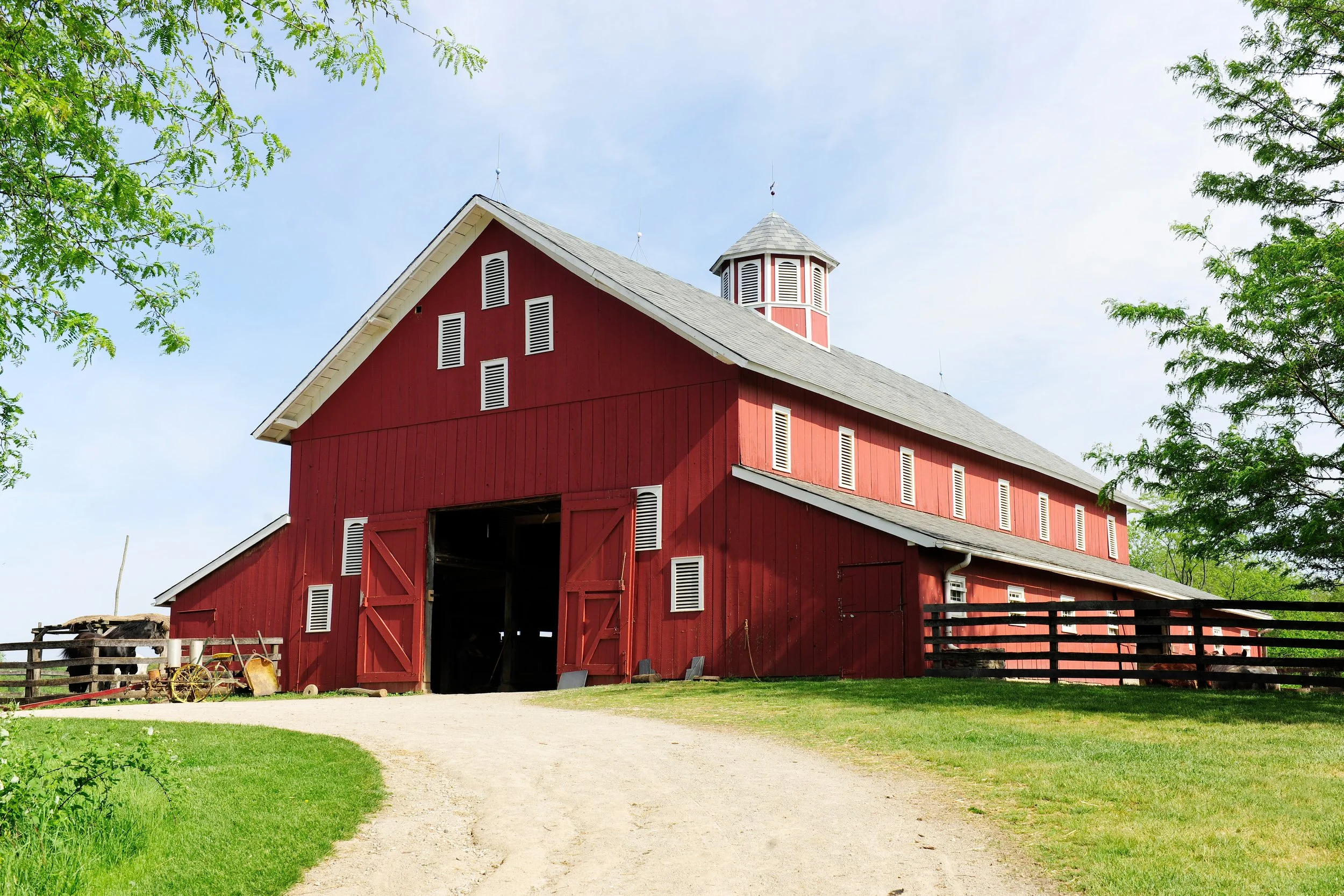 A classic red barn separated from a green lawn with a dirt path, surrounded by trees, under a partly cloudy sky.
