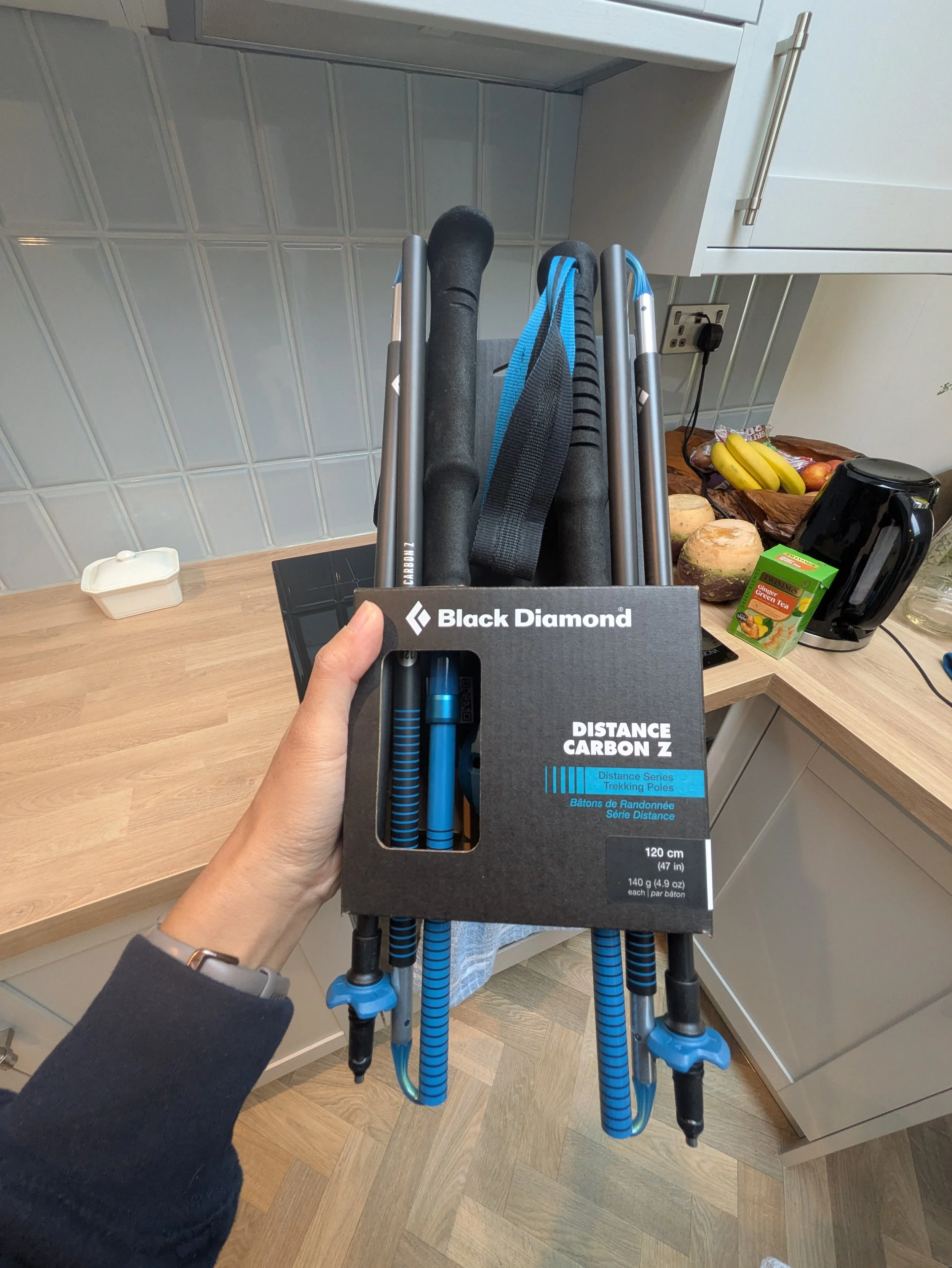 Hand holding a box of Black Diamond distance carbon trekking poles with several blue and black trekking poles attached to a cart in a kitchen.