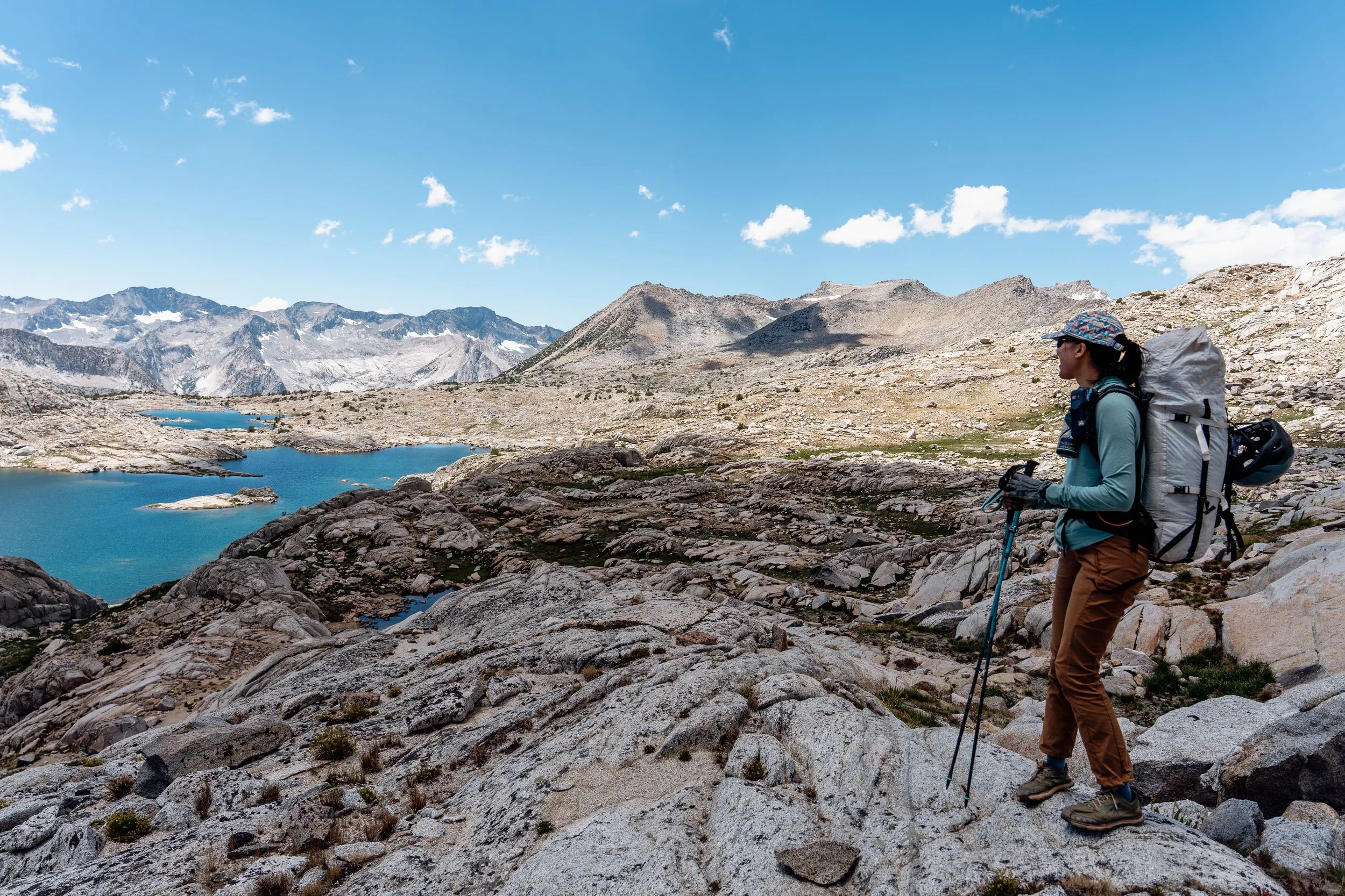 A woman hiking in a mountainous landscape with lakes, rocky terrain, and distant snow-capped peaks under a blue sky.