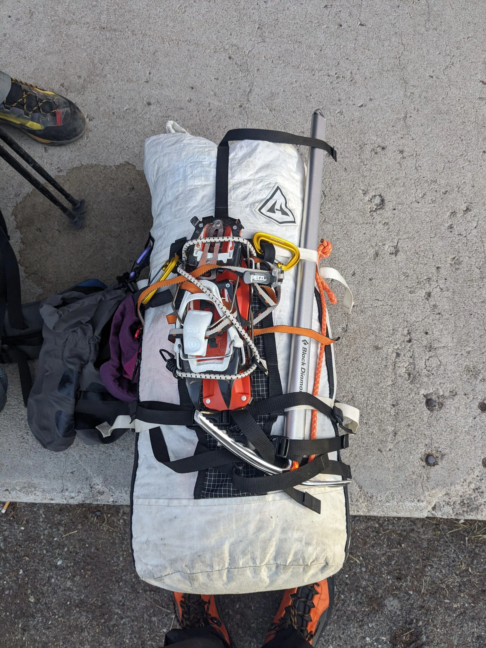 A backpack laid on a ground covered in dirt and concrete, equipped with climbing gear including carabiners, a harness, and a helmet, with a person's orange and black hiking shoes visible at the bottom of the image.