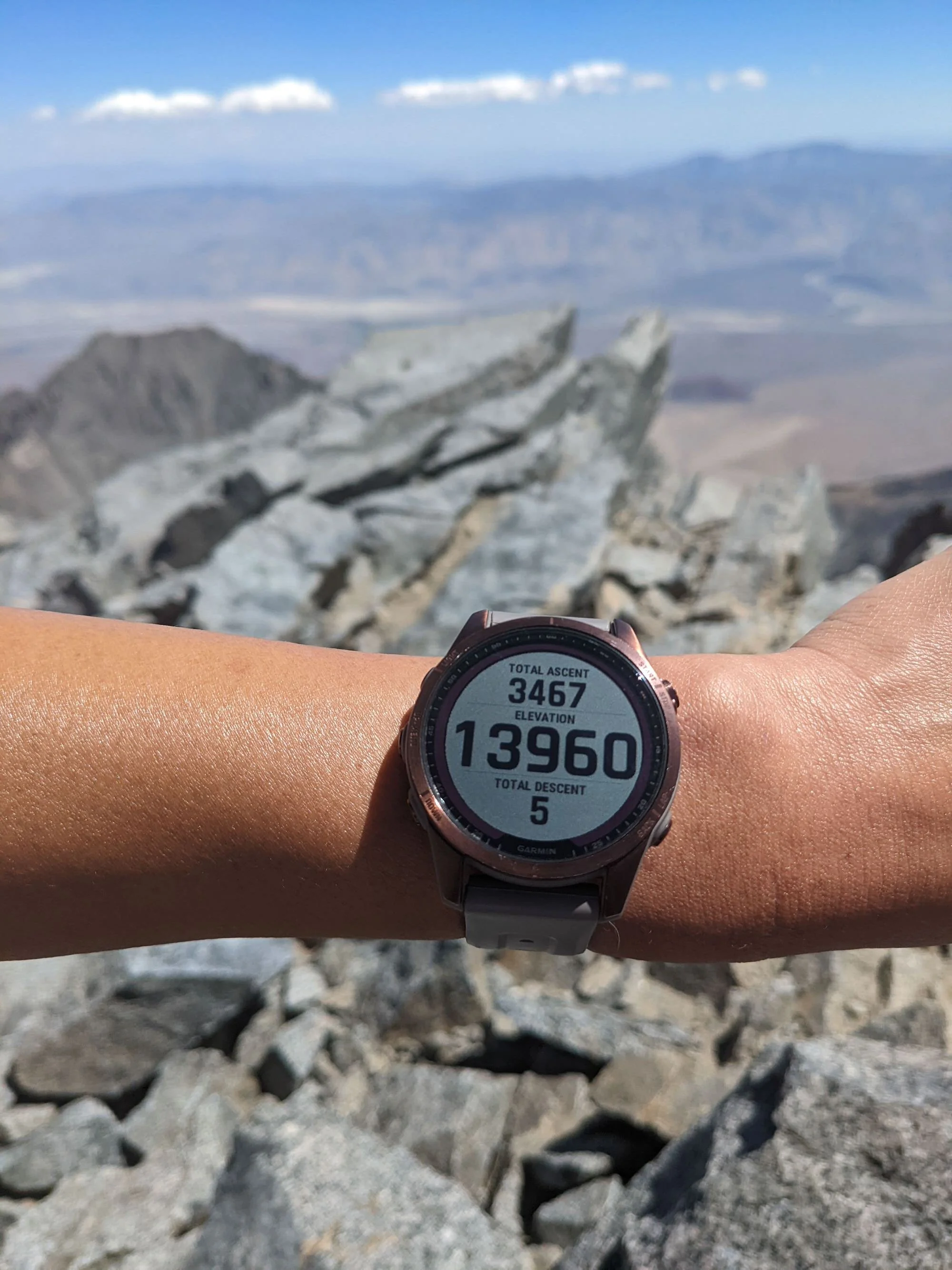 Close-up of a wrist wearing a GPS watch with a mountain landscape in the background. The watch displays total ascent of 3,467 feet, elevation of 13,960 feet, and total descent of 5 feet amidst rocky terrain.