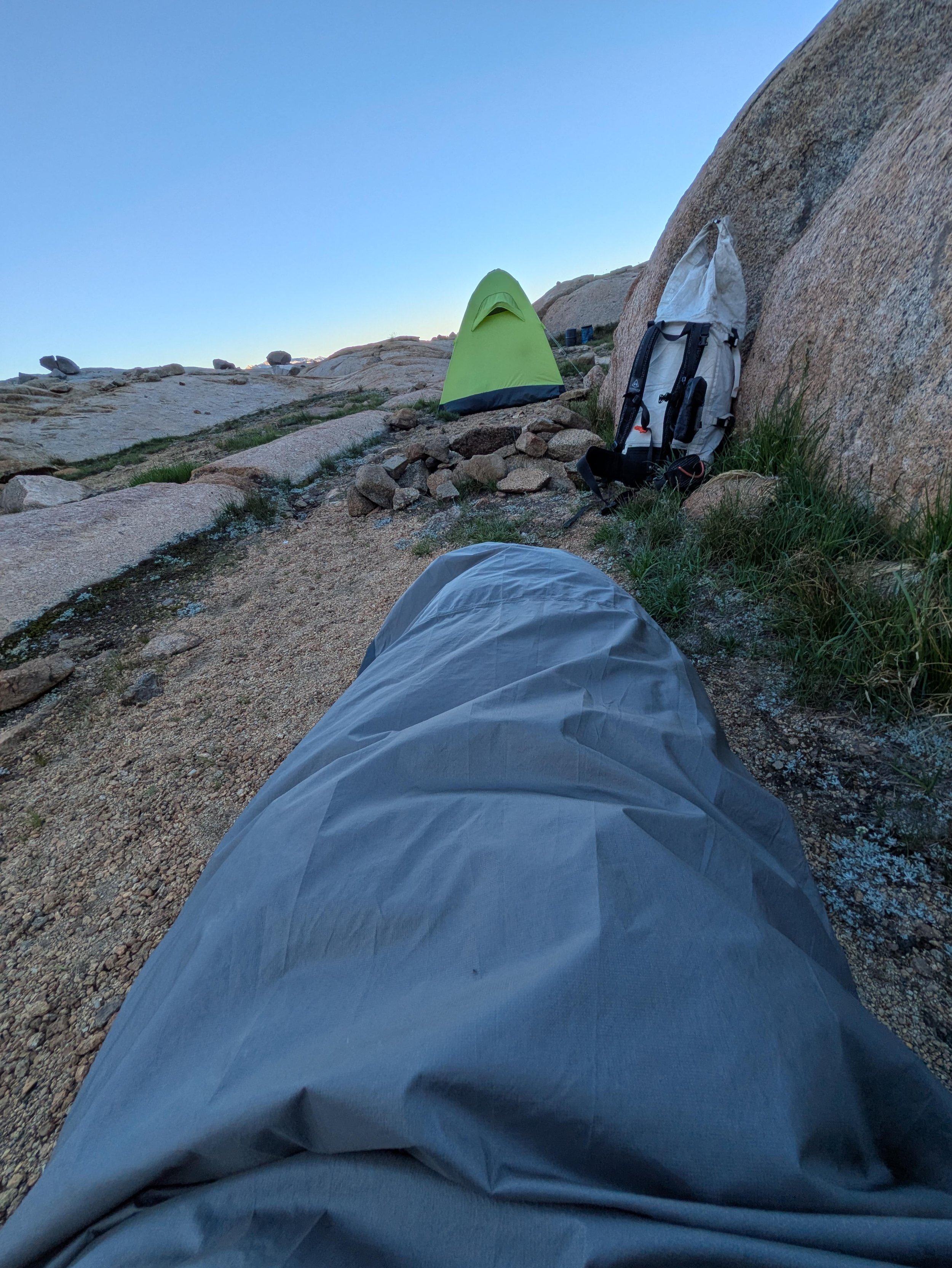 View from the ground of a camping site with a yellow tent, a gray backpack leaning against a large rock, and gravel and grass on the ground under a clear blue sky.