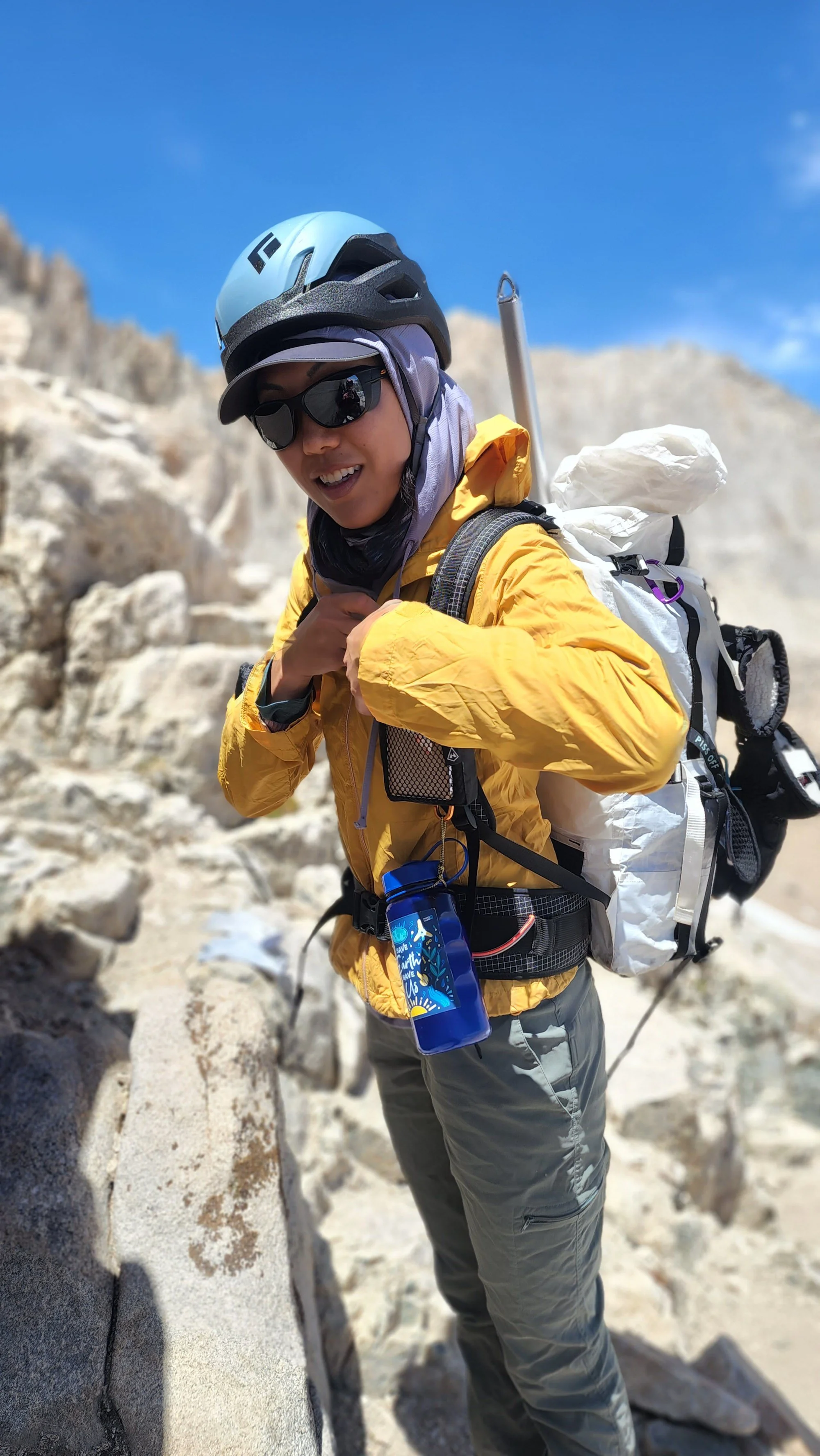 Person wearing a yellow jacket, sunglasses, and a helmet climbing rocky terrain with a backpack in a mountainous area.