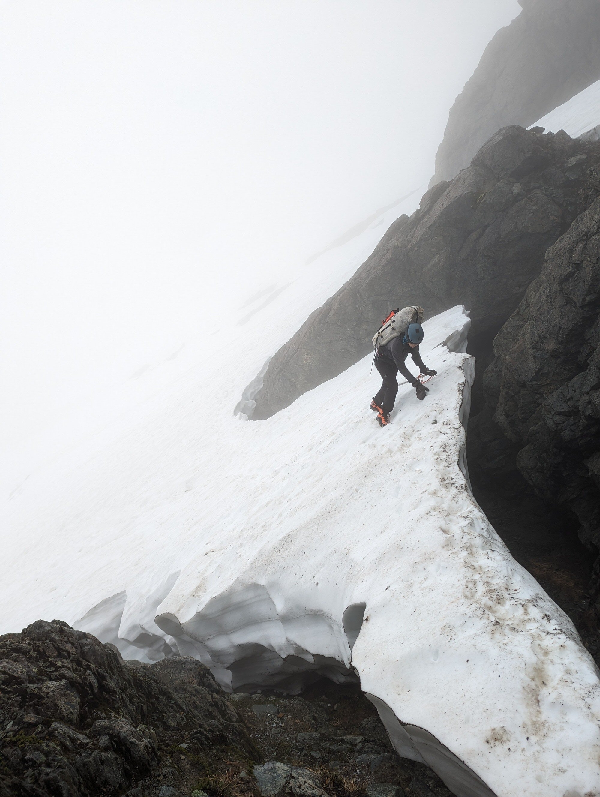 A person wearing a helmet and climbing gear is ascending a snow-covered mountain slope, holding an ice axe, surrounded by large rocks and surrounded by fog.