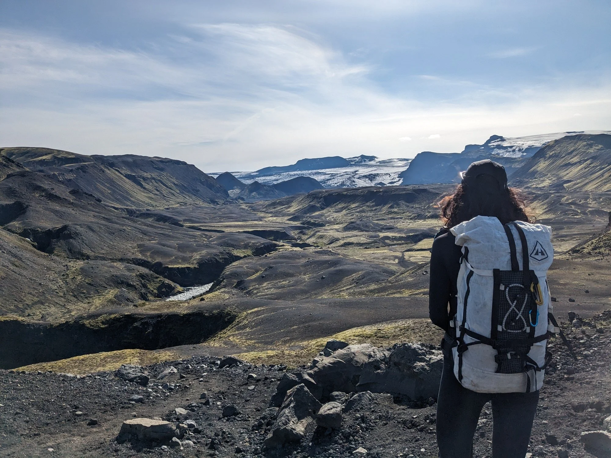 Person with curly hair wearing a cap and a white hiking jacket, carrying a backpack, hiking in a rugged volcanic landscape with mountains and patches of snow under a partly cloudy sky.