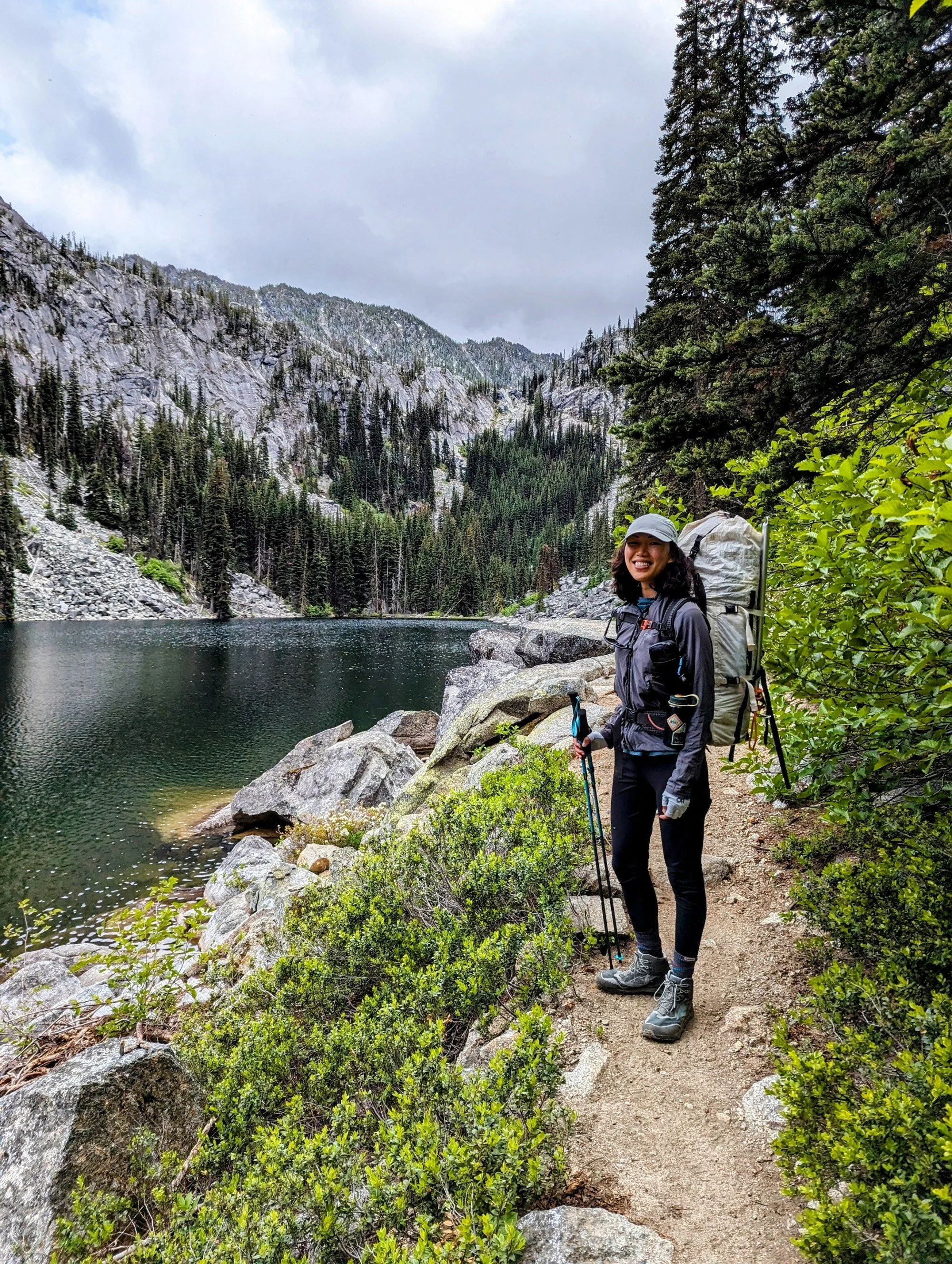 A smiling woman hiking along a trail next to a lake in a mountainous forest, carrying a large backpack and using trekking poles.