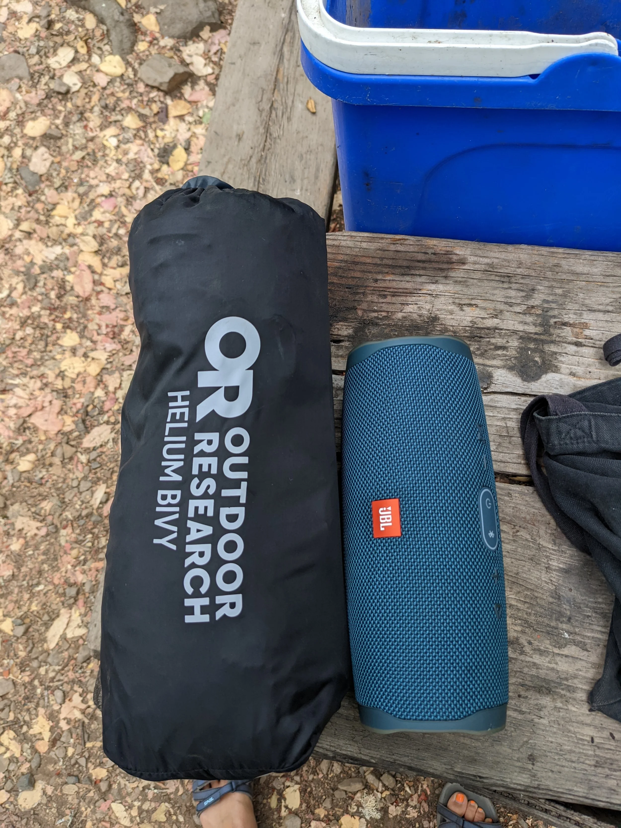 Arrangement of outdoor items on a wooden bench, including a black bag with white text, a blue portable JBL speaker, a pair of black sandals, and a black hat. The ground is covered with leaves and small rocks.