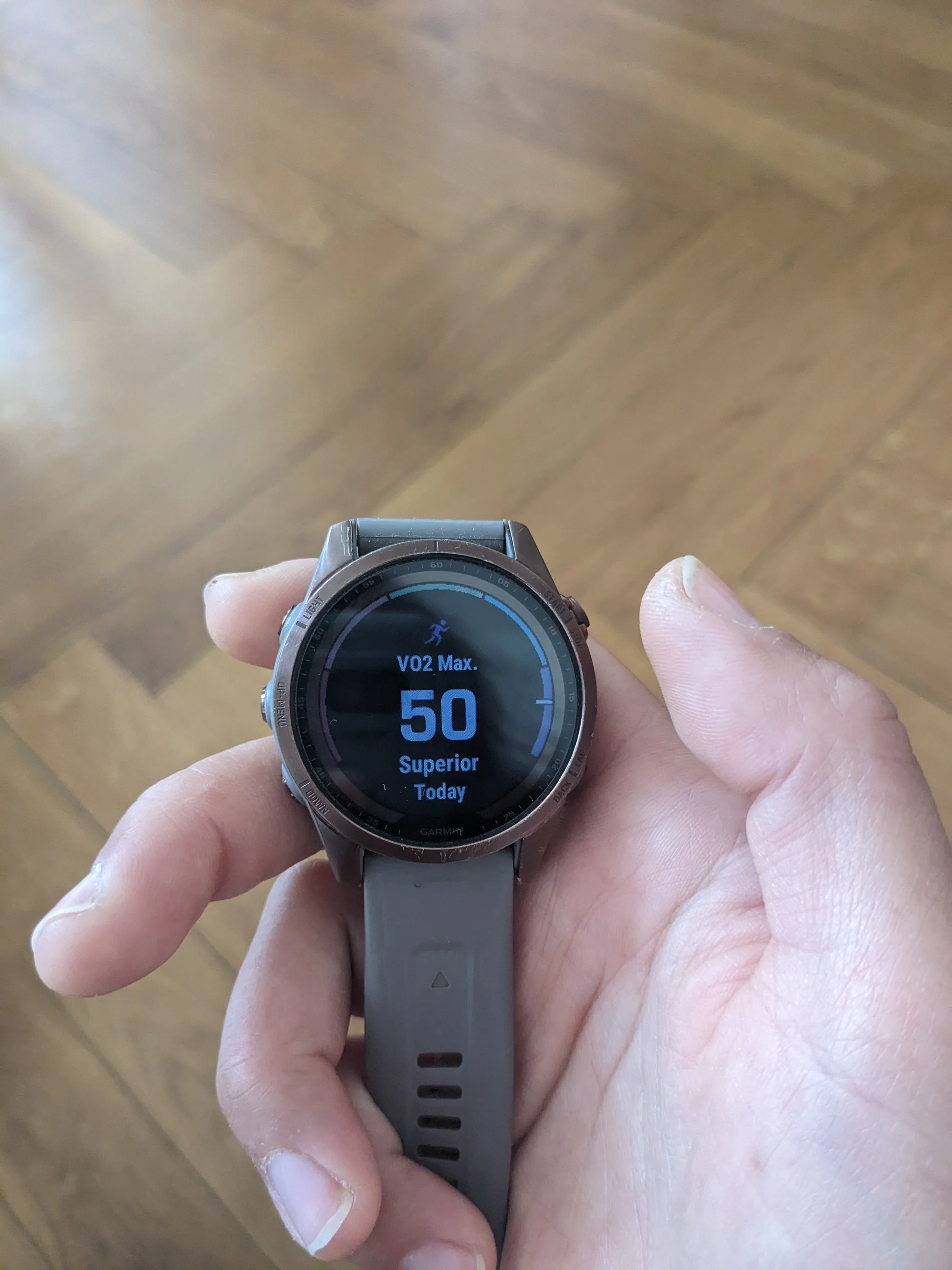 A person holding a smartwatch displaying a VO2 max of 50, with a background of a wooden floor.