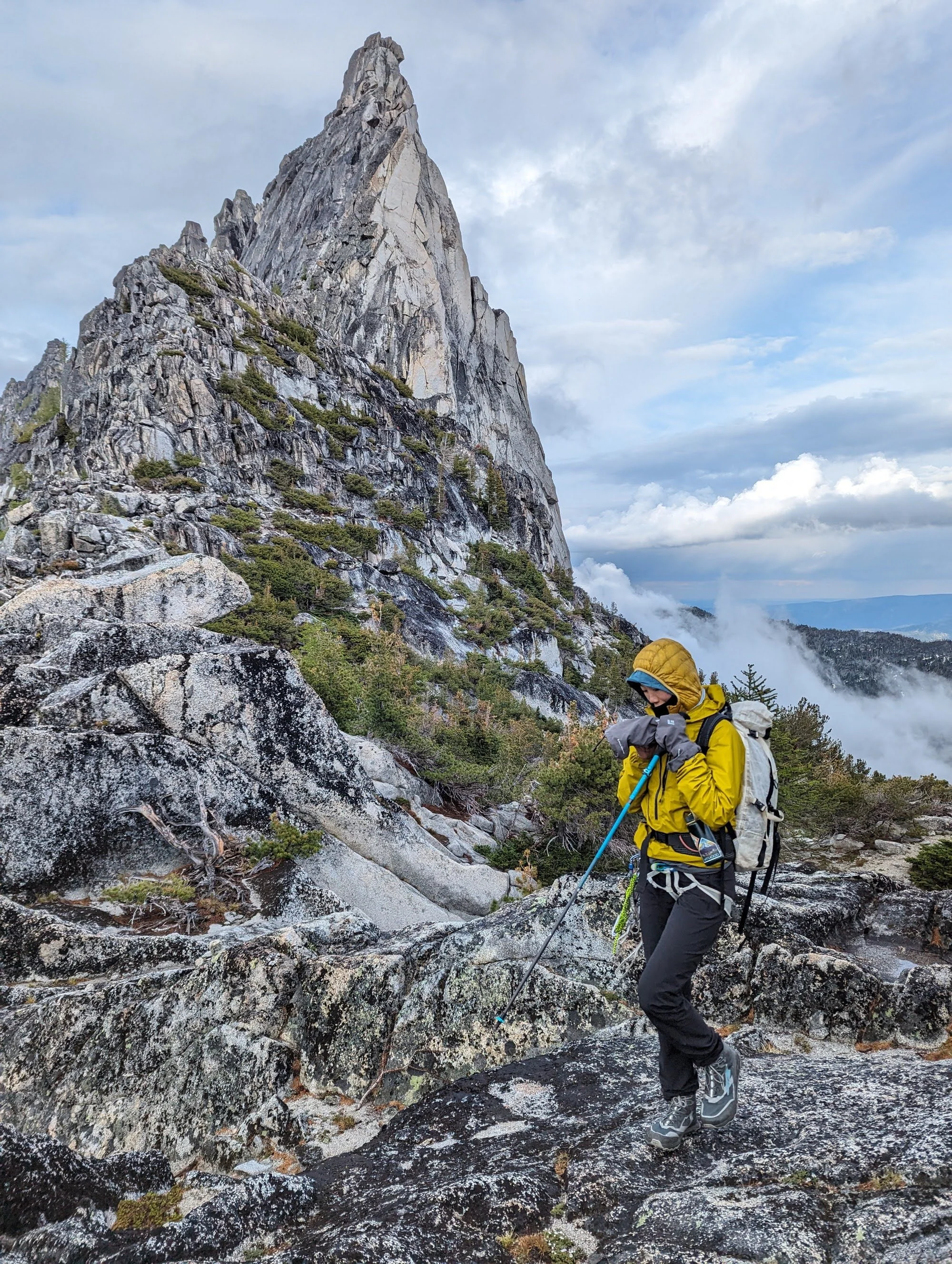 A person wearing a yellow rain jacket and hiking boots uses hiking poles on rocky terrain with a mountainous background.