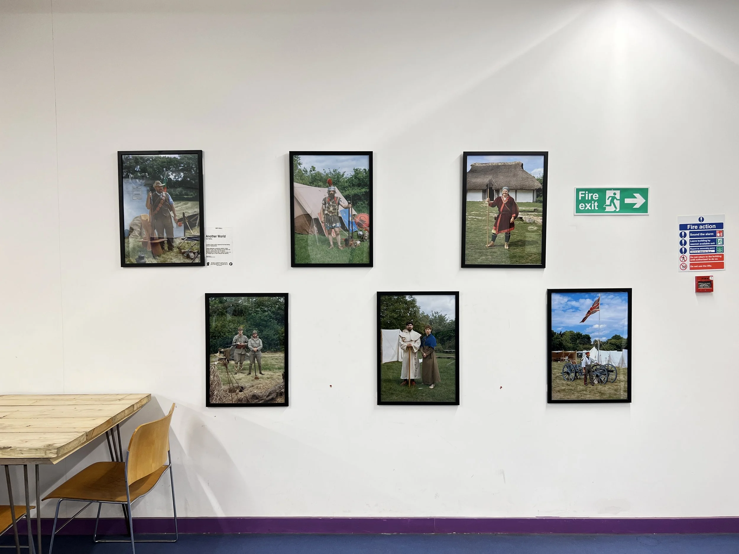 Gallery wall with six framed photographs depicting historical reenactments and people in period costumes, with a fire exit sign to the right and a fire action notice, in an indoor setting.