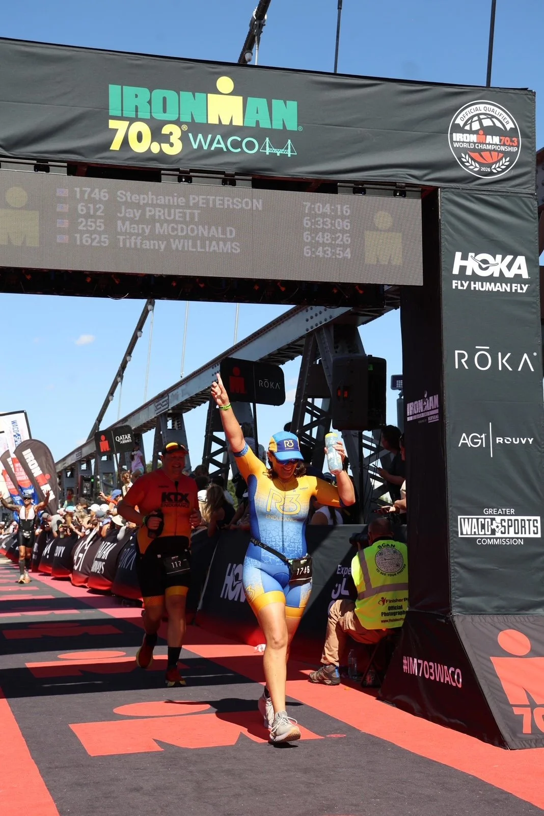 Female athlete crossing the finish line of an Ironman race, wearing a blue and yellow triathlon suit, holding a water bottle, with a crowd and race officials in the background, and race information displayed on a large screen above.