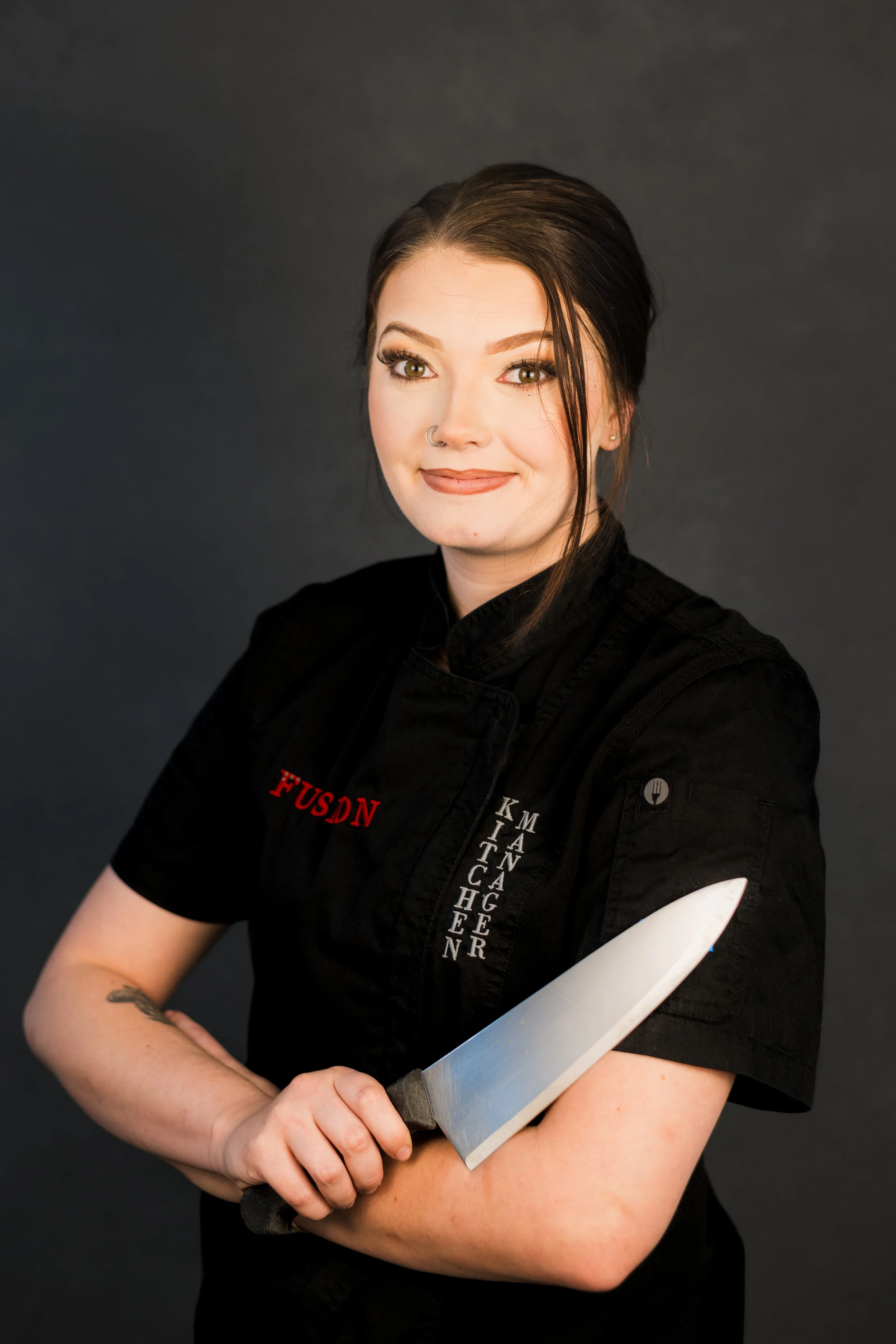 Yevette Lynn, brand photographer, creates a studio chef portrait featuring a woman in a black uniform holding a knife across her chest.