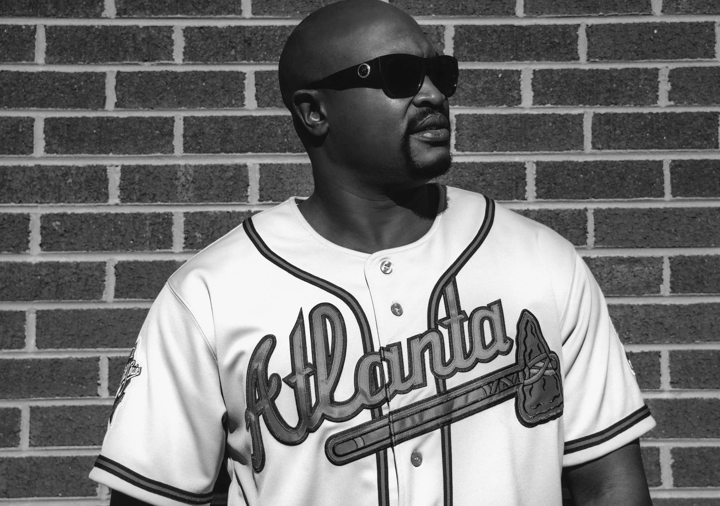 Yevette Lynn portrait photographer captures black and white outdoor portrait of man wearing Atlanta baseball jersey and sunglasses against brick wall.
