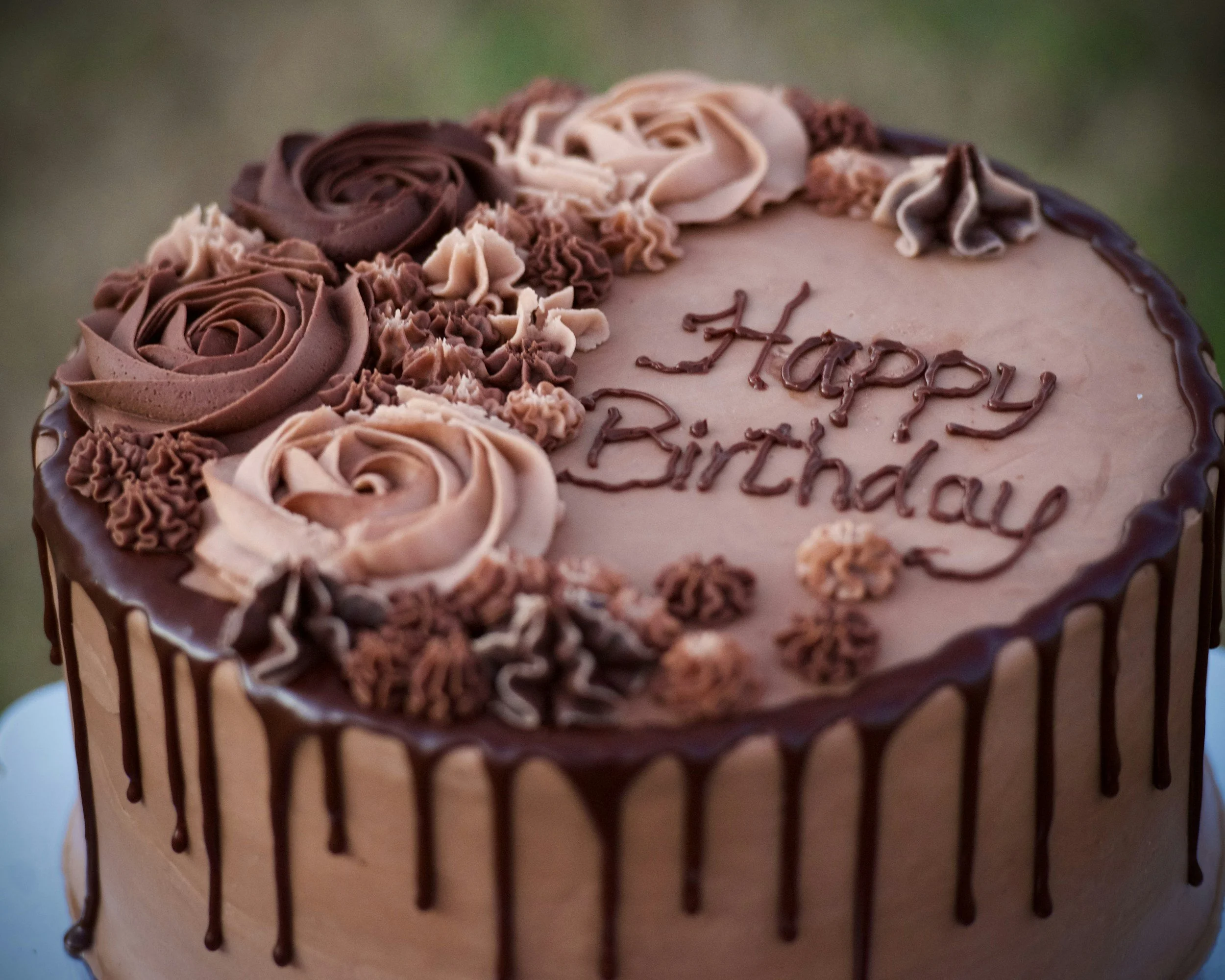 A birthday cake decorated with pink and brown frosting, with a drizzle of chocolate on the sides and piped roses and flowers on top, with the message "Happy Birthday" written in chocolate.