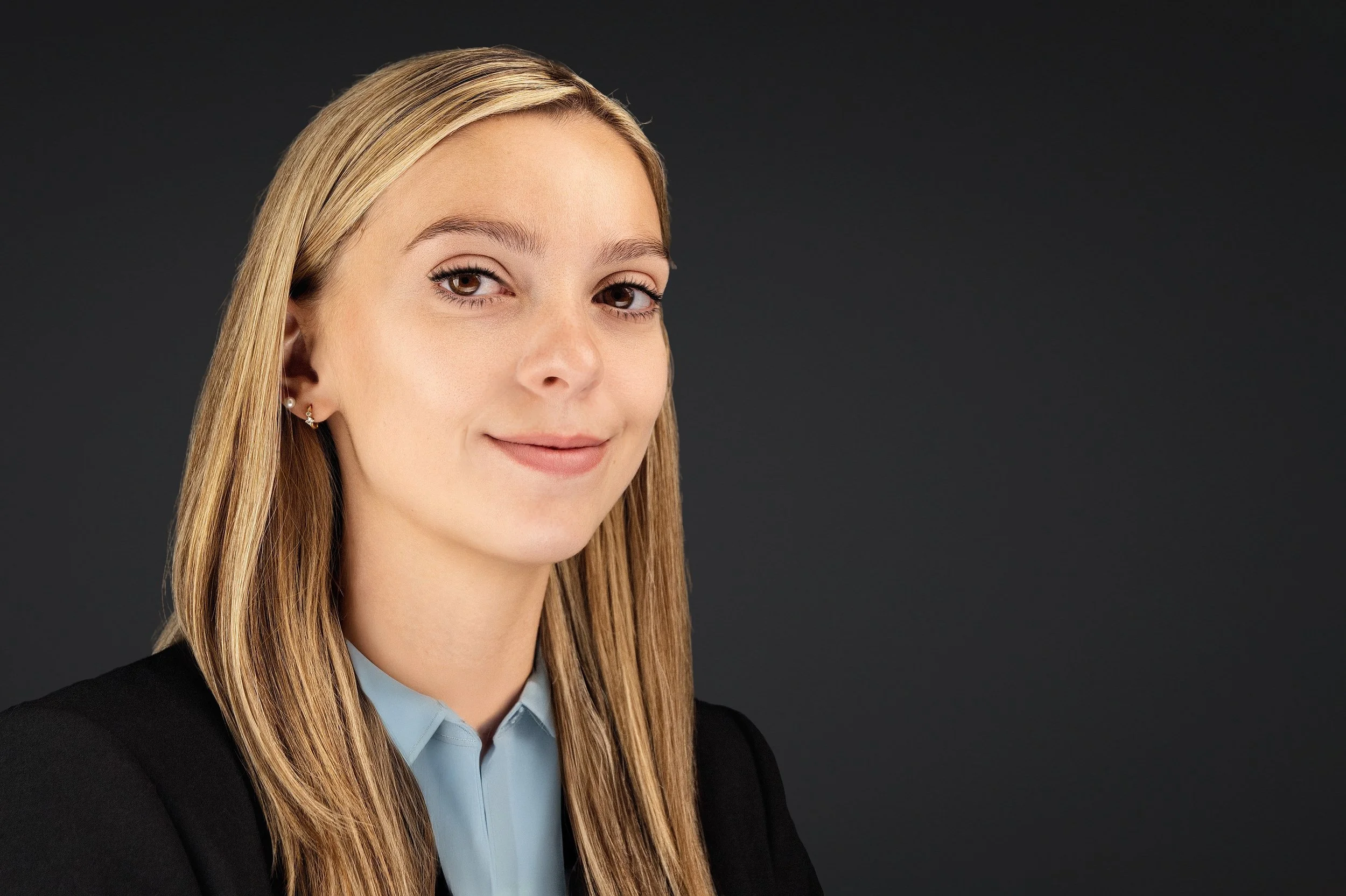 Yevette Lynn, professional photographer, creates a refined studio headshot of a blonde woman in a blazer with a soft smile against a dark background.