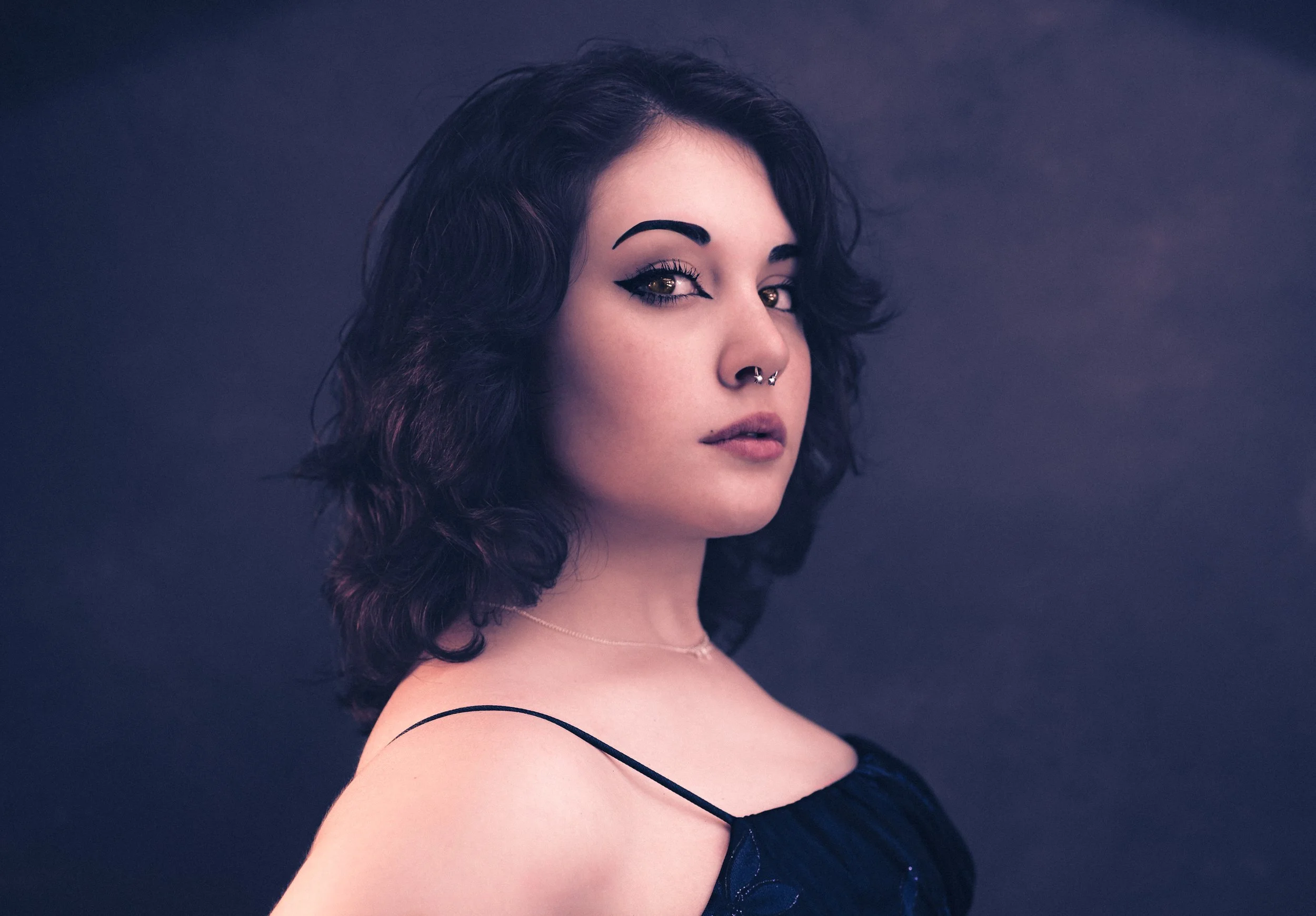 Yevette Lynn portrait photographer captures studio portrait of woman with dark wavy hair and nose ring, posed in profile against moody background.