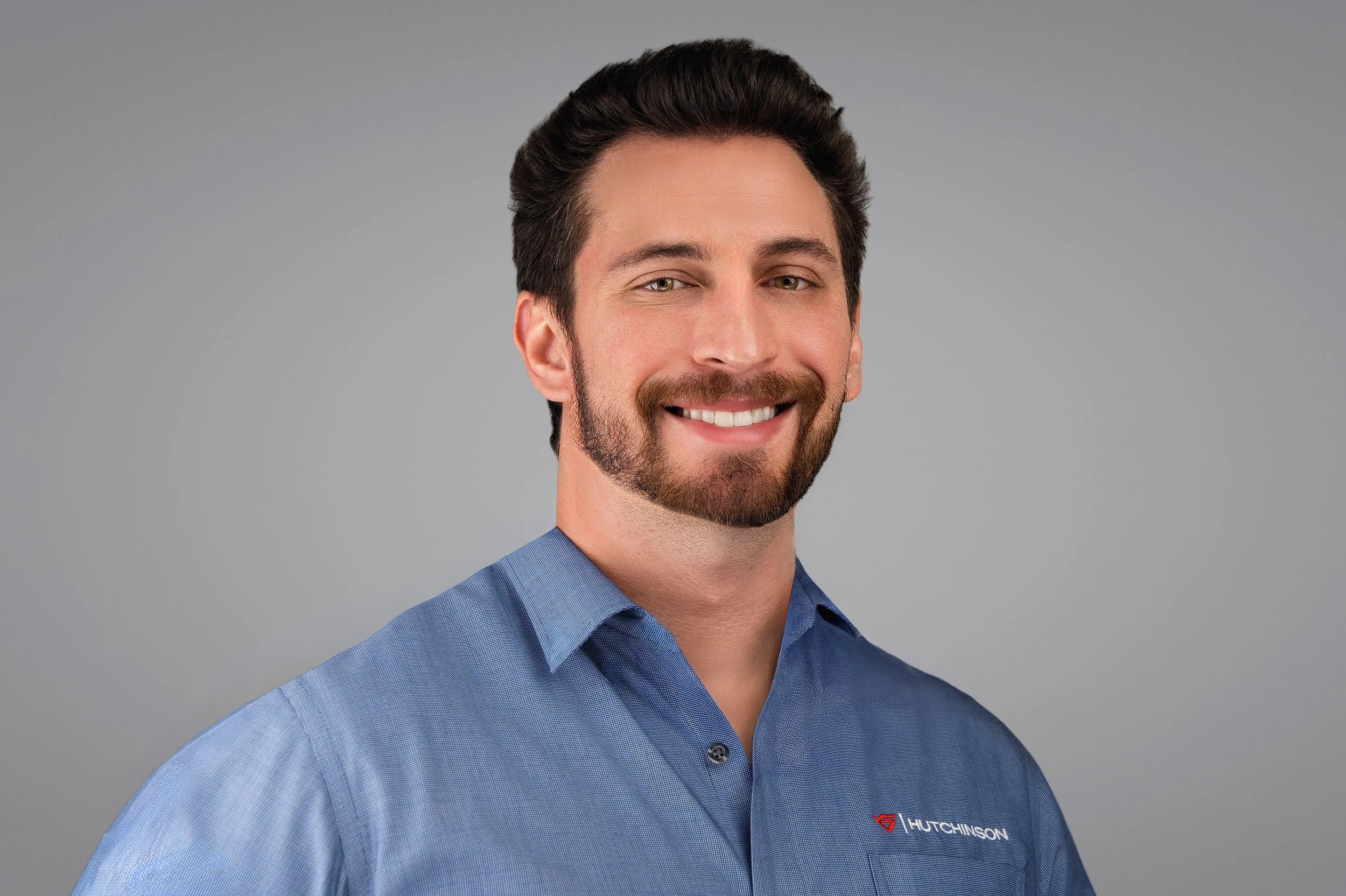 Yevette Lynn, professional photographer, captures a friendly studio headshot of a man with a short beard wearing a blue button-down shirt against a gray background.