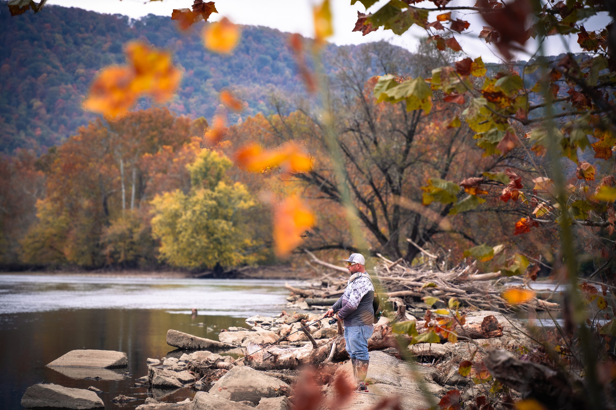 A person fishing on a rocky shoreline beside a river, framed by autumn-colored fall leaves and trees with mountain in the background.