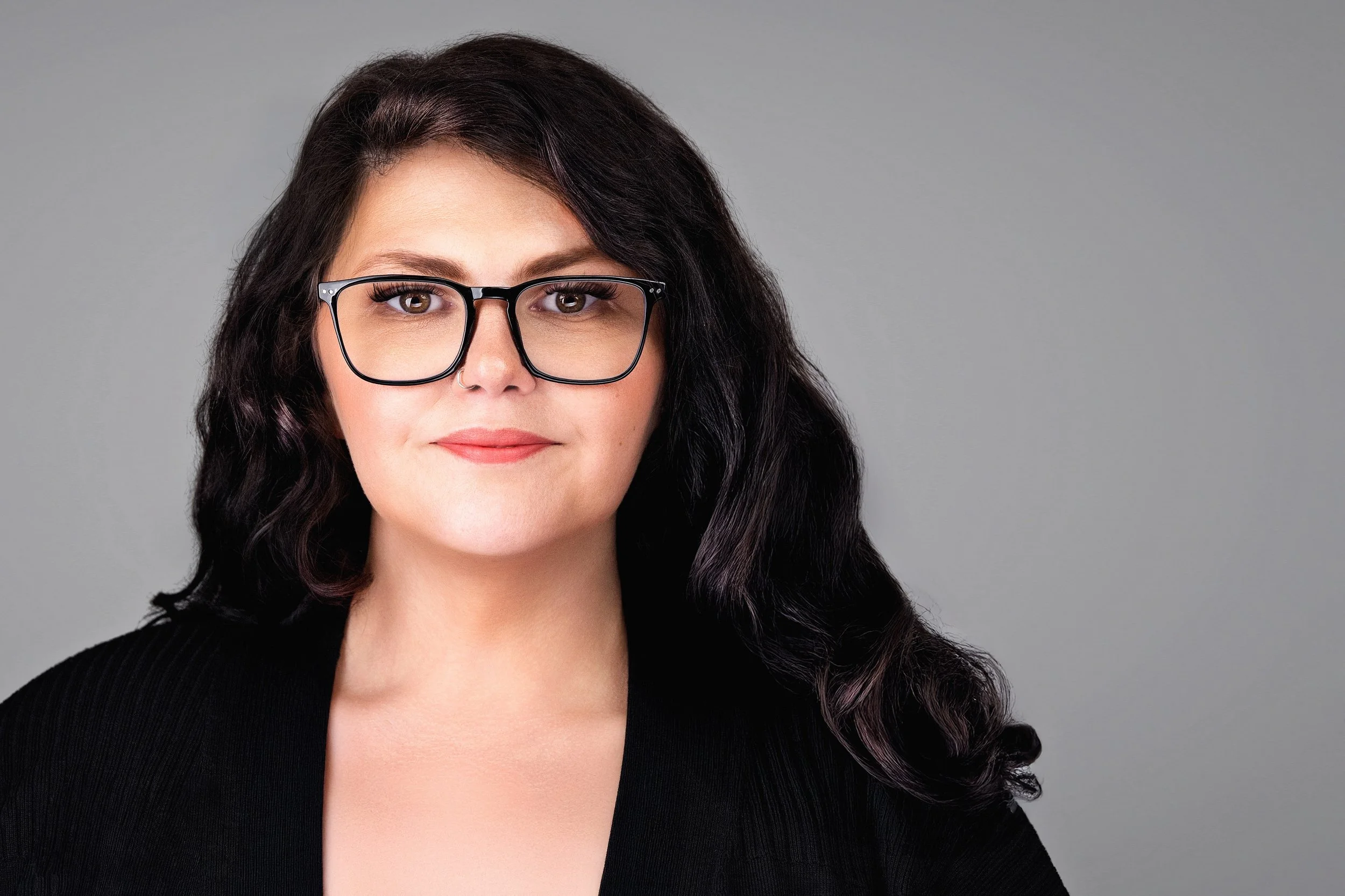 Yevette Lynn, professional photographer, captures a polished studio headshot of a woman wearing glasses and a black blazer against a soft gray background.