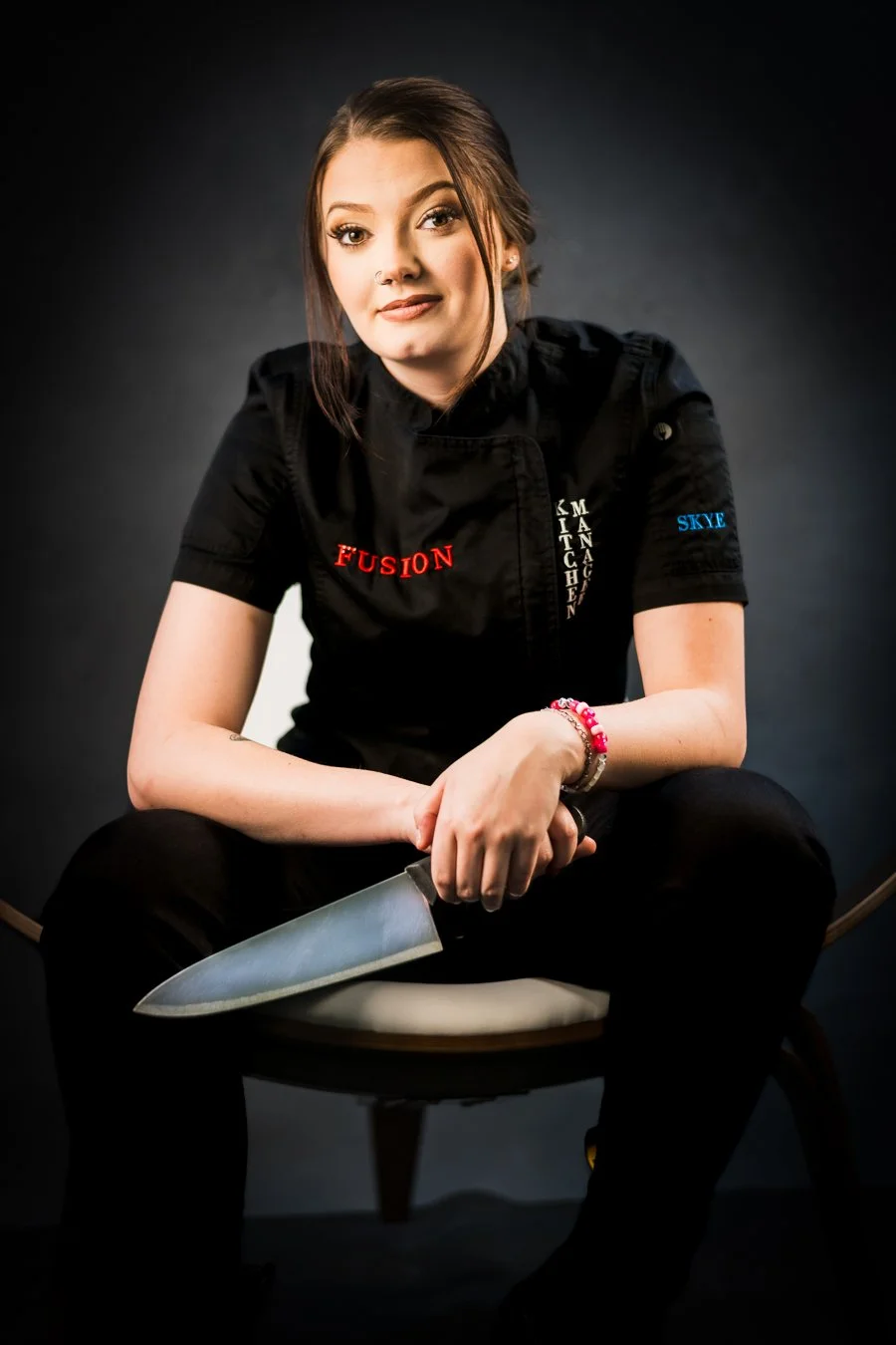 Yevette Lynn, brand photographer, captures a studio portrait of a chef seated holding a knife with calm confidence against a dark background.