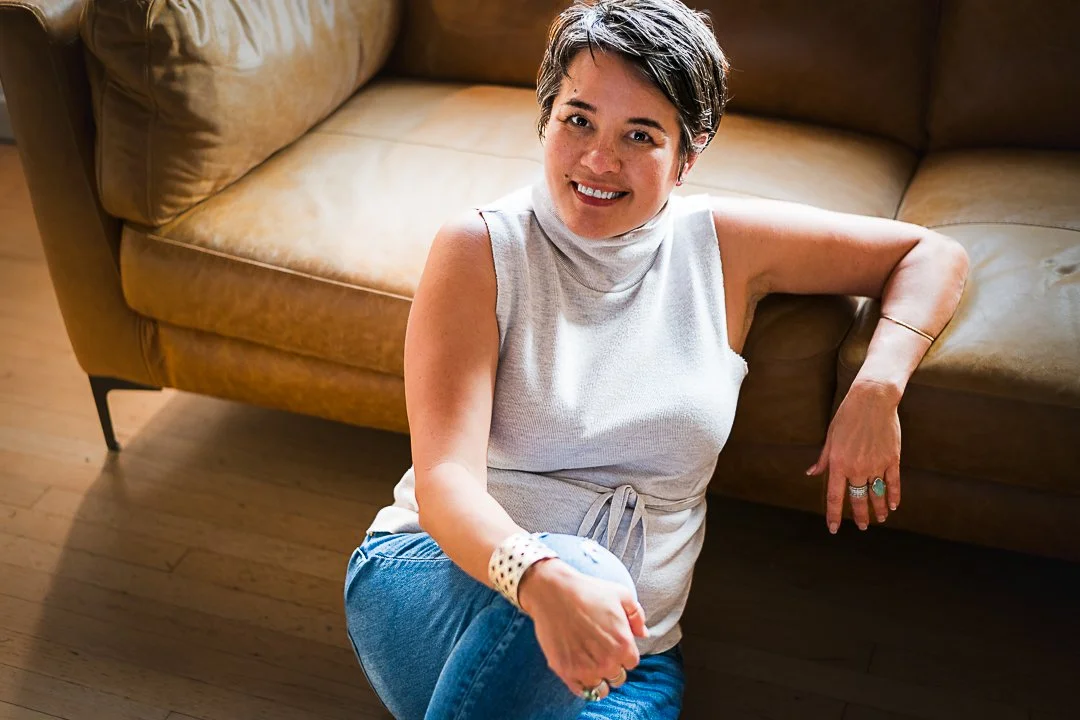 Yevette Lynn portrait photographer captures lifestyle portrait of woman sitting on floor beside sofa, smiling naturally in softly lit home interior.
