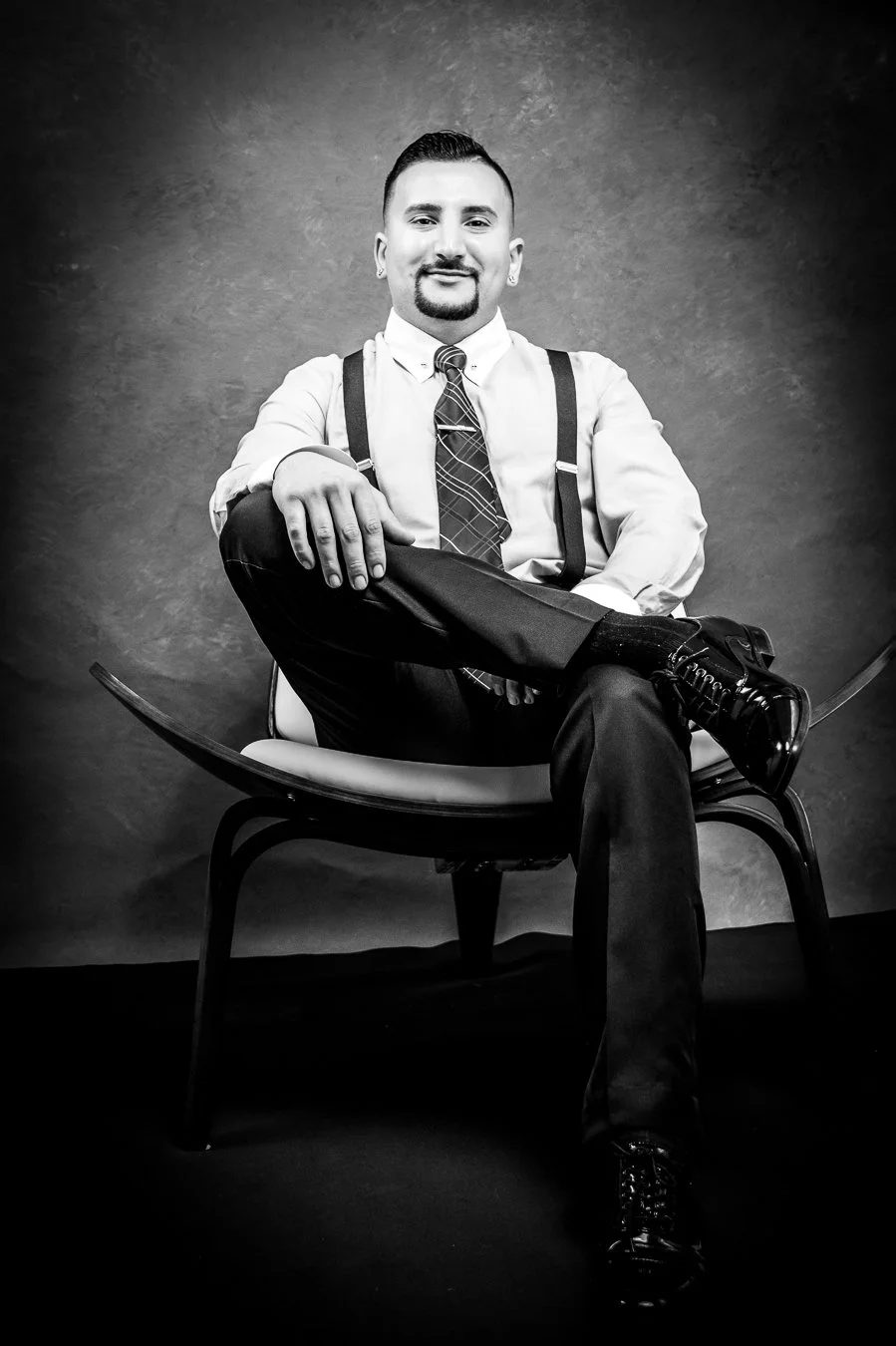 Yevette Lynn portrait photographer creates black and white professional portrait of seated man wearing suspenders and tie, relaxed pose in studio setting.