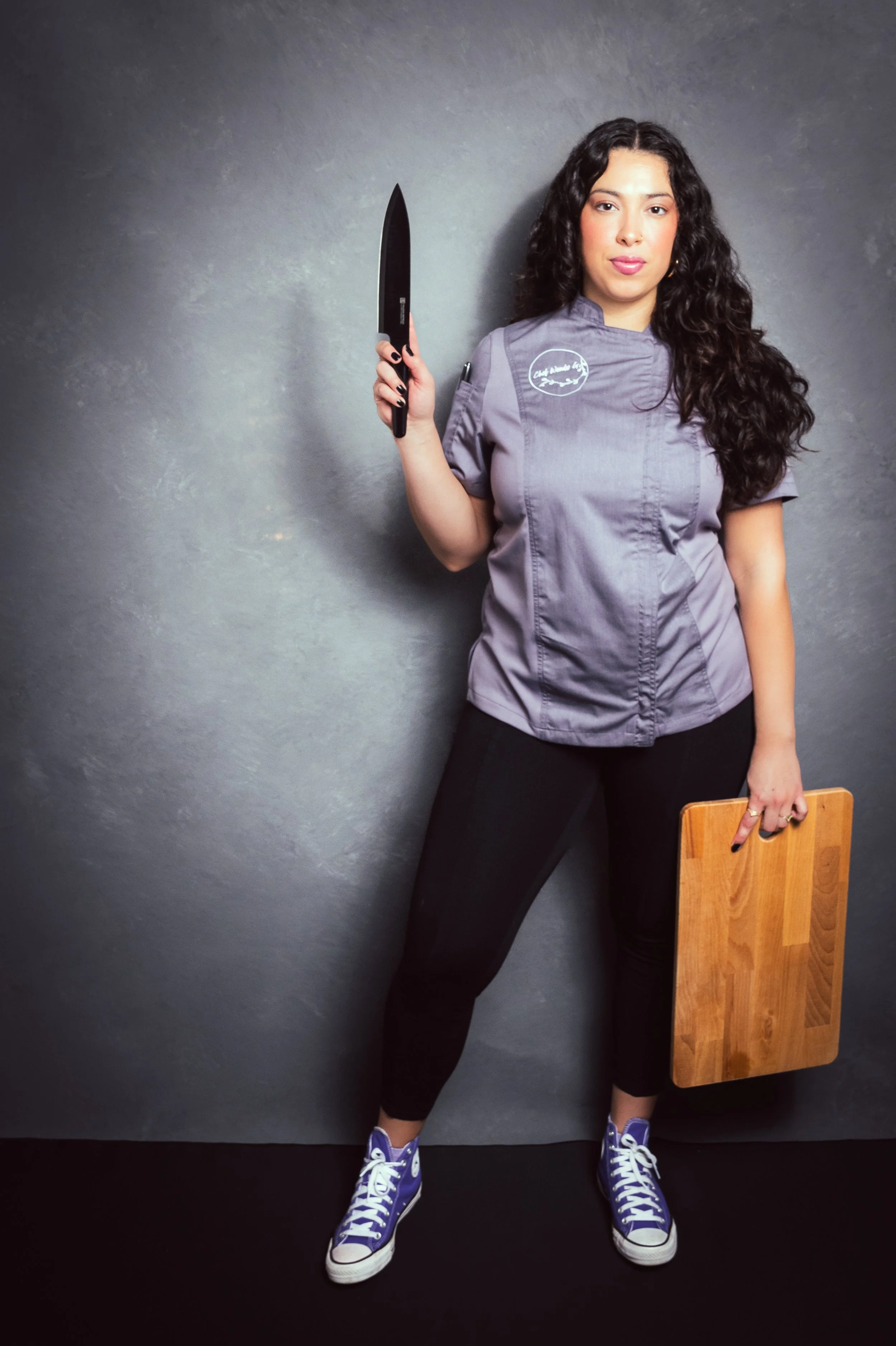 Yevette Lynn, brand photographer, captures a studio brand portrait of a woman chef in a gray uniform holding a knife and cutting board against a textured backdrop.