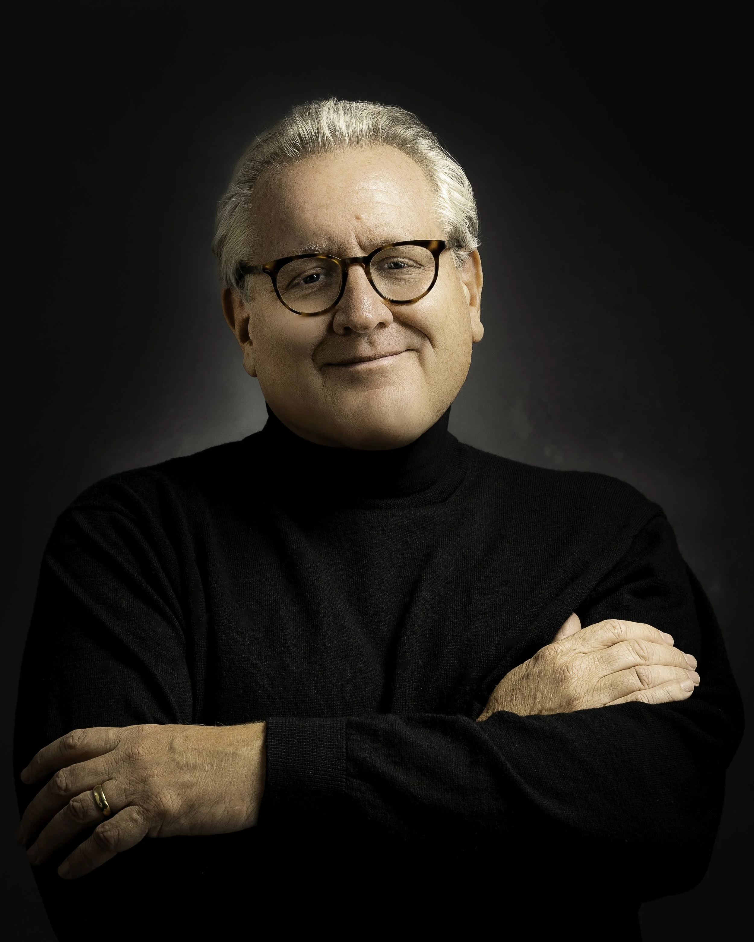 Yevette Lynn portrait photographer captures professional studio portrait of an older man wearing glasses and black sweater, arms crossed against dark background.
