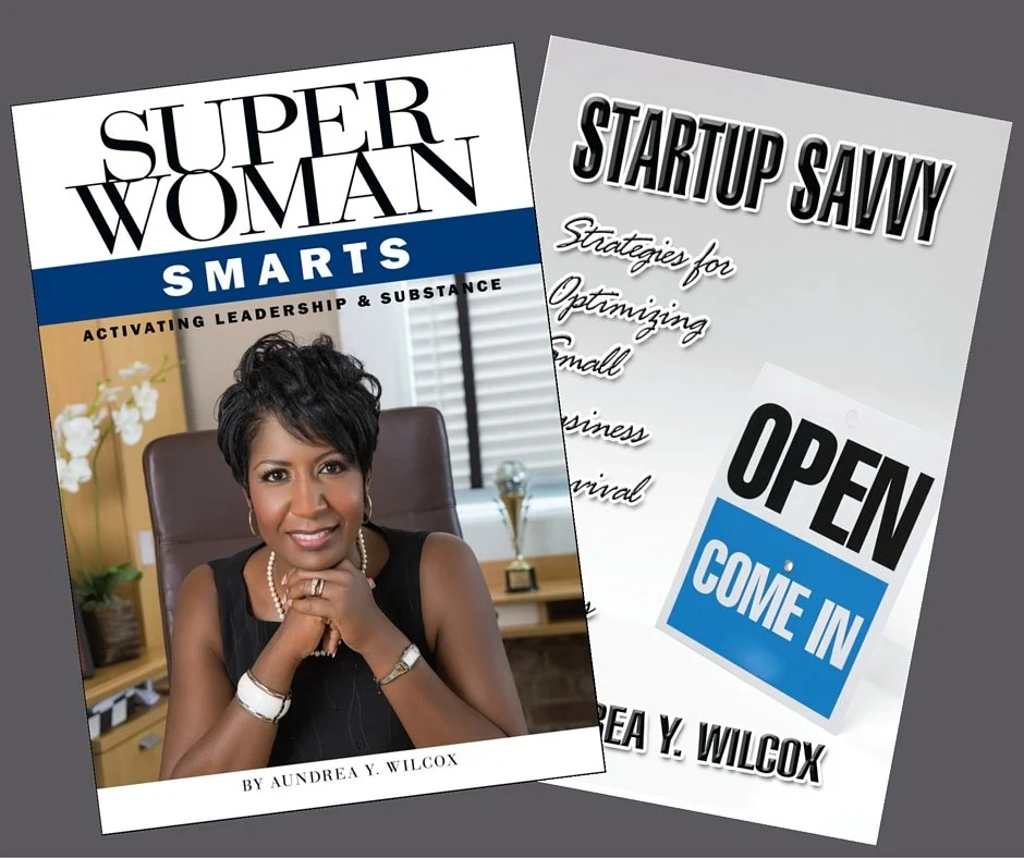Two books written by Yevette Lynn, one titled "Super Woman Smarts" featuring Yevette Lynn with short black hair sitting in an office chair, and the other titled "Startup Savvy" with an "Open Come In" sign, on a gray surface.
