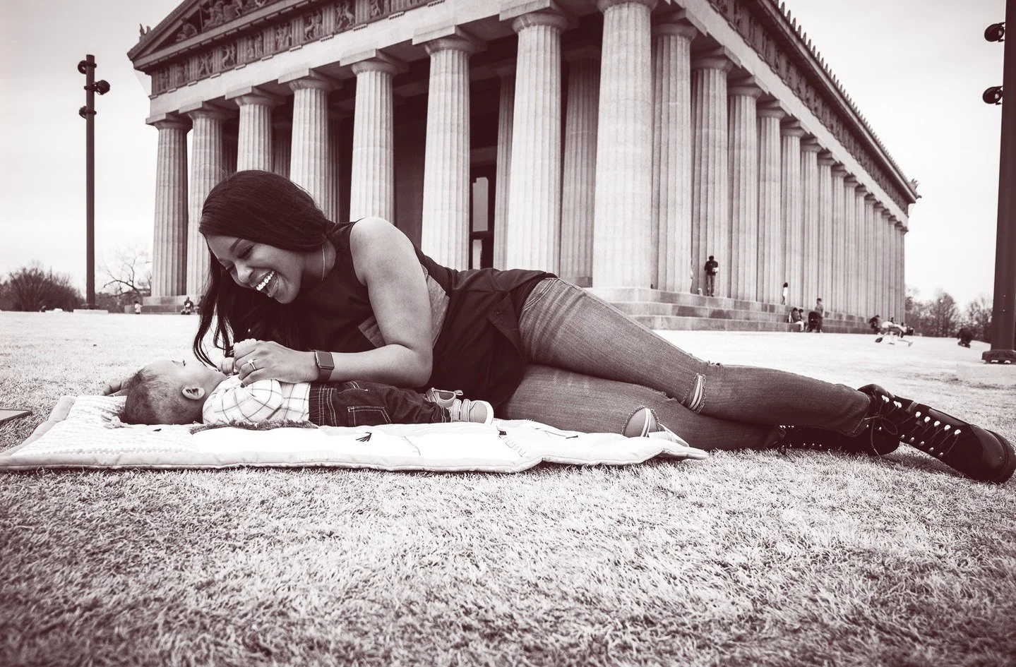 Yevette Lynn, lifestyle photographer, creates a black and white outdoor portrait of a mother lying on a blanket with her baby near classical columns.