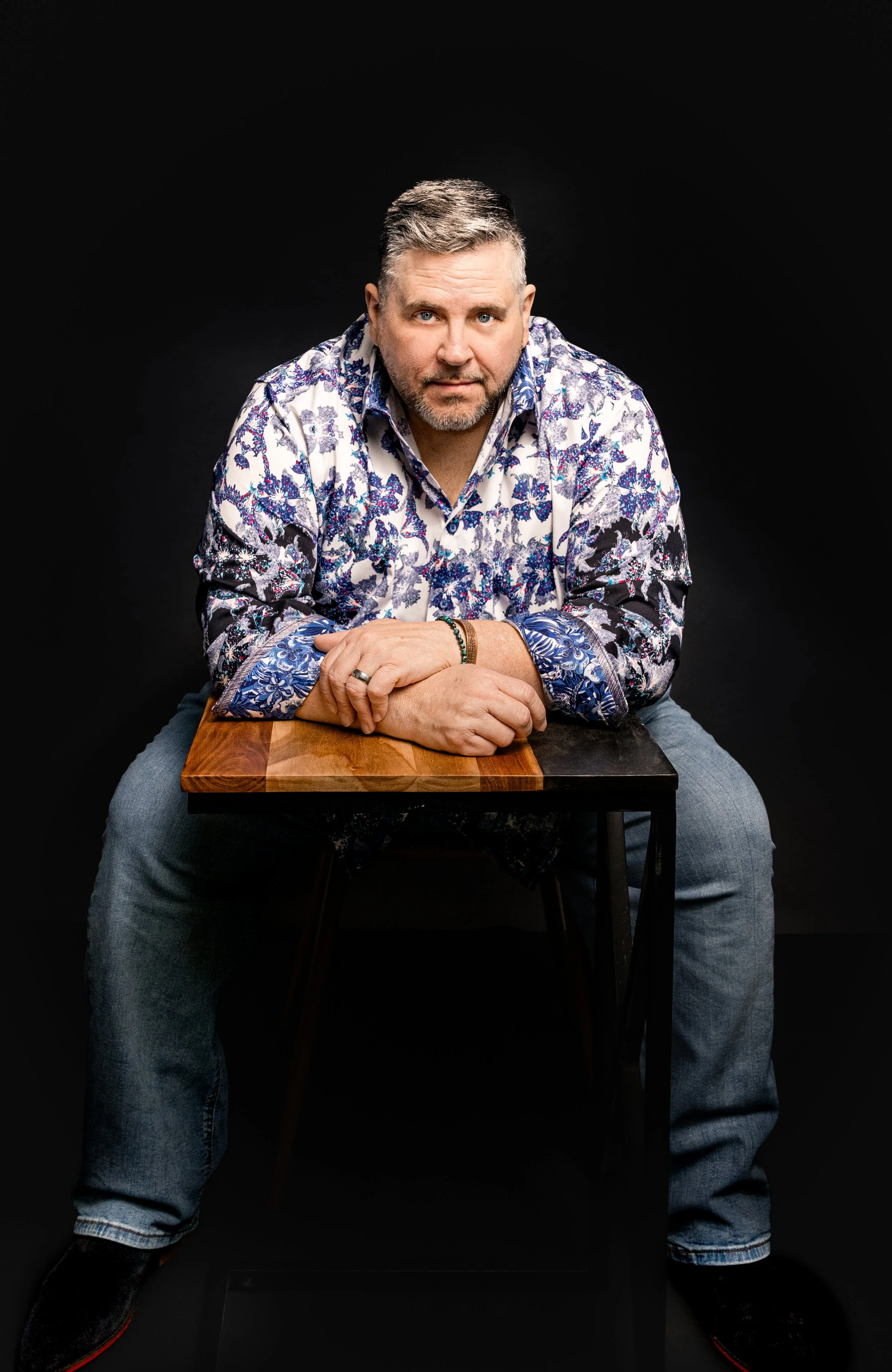 Yevette Lynn portrait photographer captures studio portrait of a seated man leaning forward at table, wearing patterned shirt against dark background.