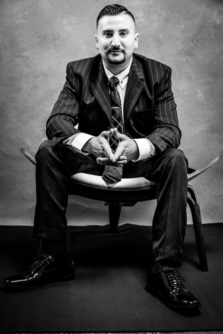 Yevette Lynn portrait photographer creates black and white professional portrait of seated man in tailored suit, hands clasped, studio backdrop.