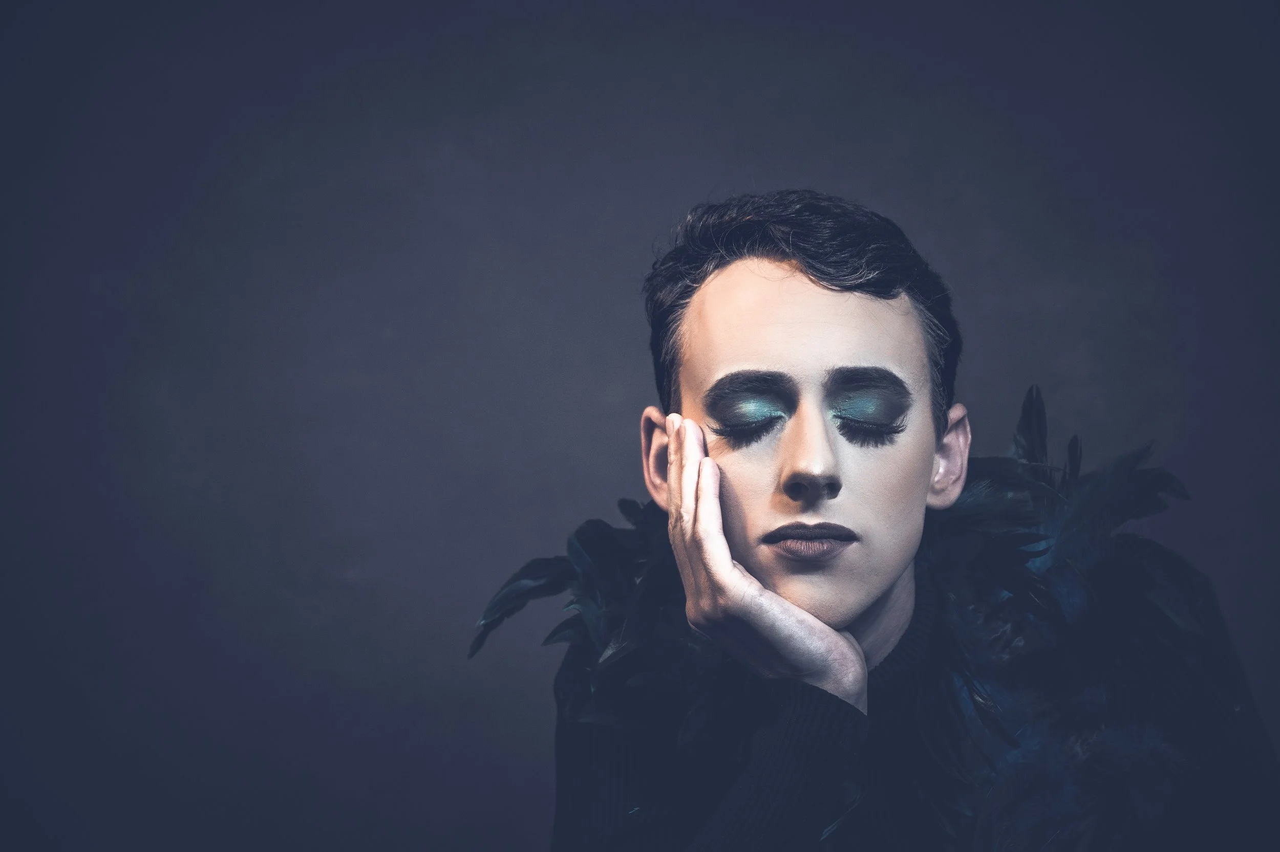 Yevette Lynn, brand photographer, captures a moody studio portrait of a man in dark fashion with eyes closed and feathered details against a dramatic background.