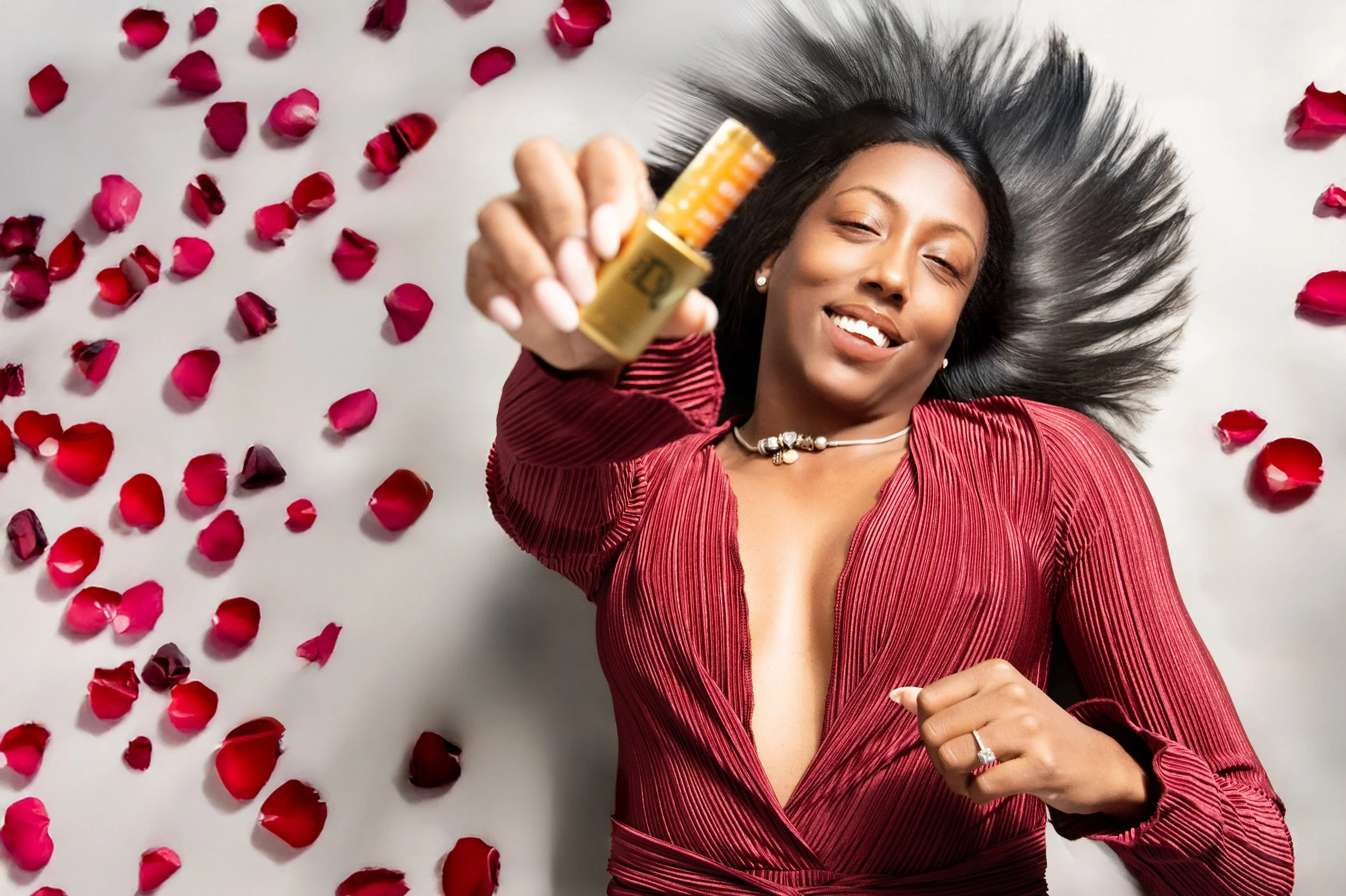 Yevette Lynn brand photographer captures beauty product photography of a woman lying among rose petals, holding skincare bottle in studio setting.