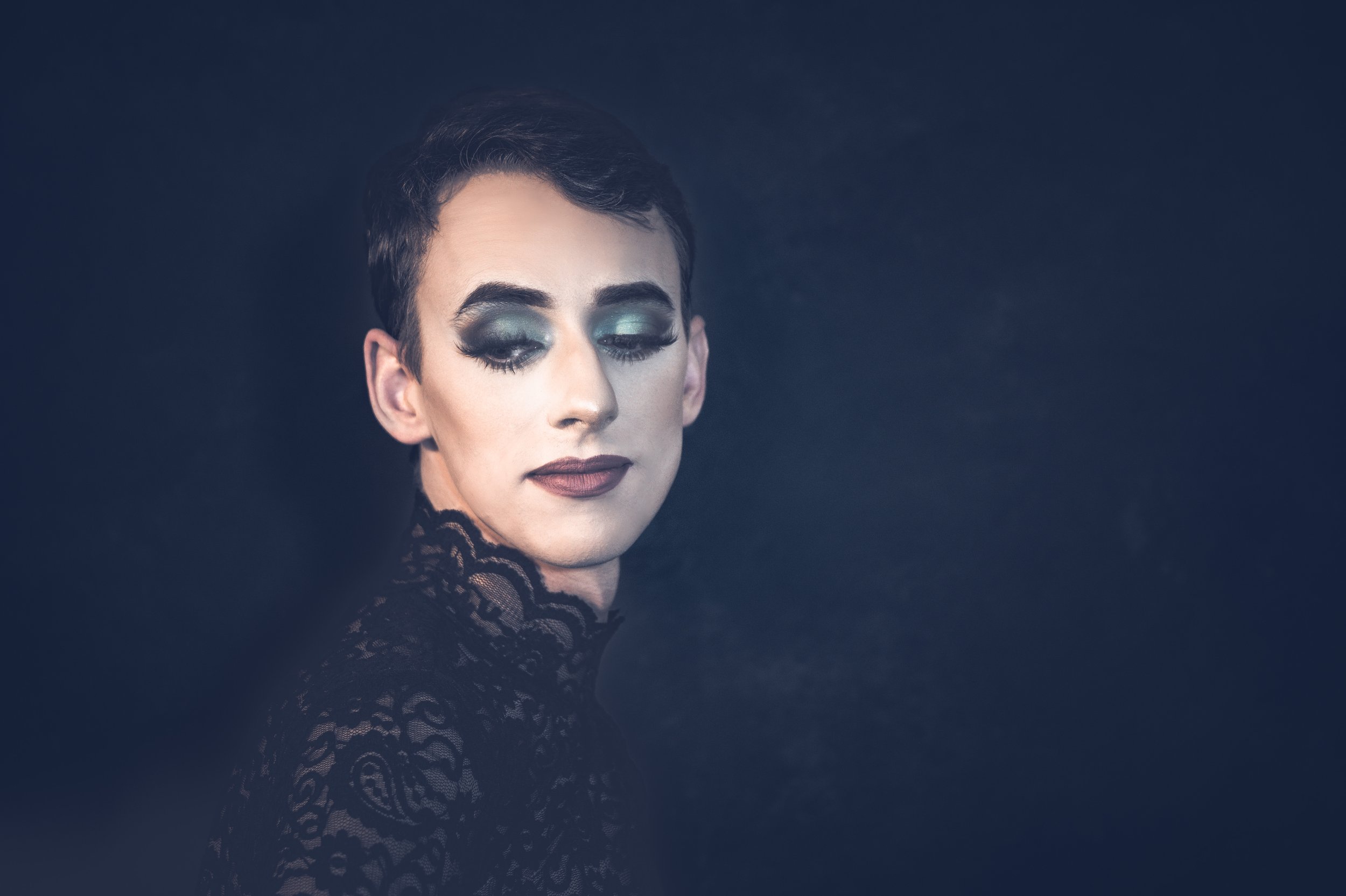 Yevette Lynn, brand photographer, captures a dramatic studio portrait of a man in dark lace fashion with bold makeup, posed in profile against a moody background.
