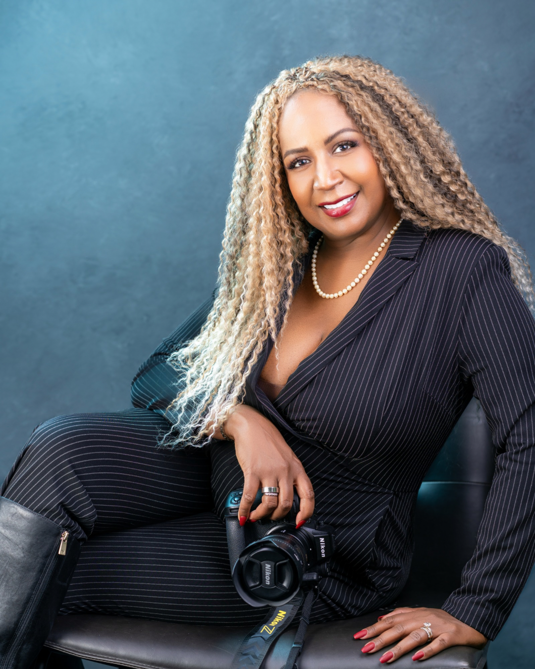 Yevette Lynn portrait photographer poses for studio headshot holding camera, seated confidently with braided hair and soft textured backdrop.