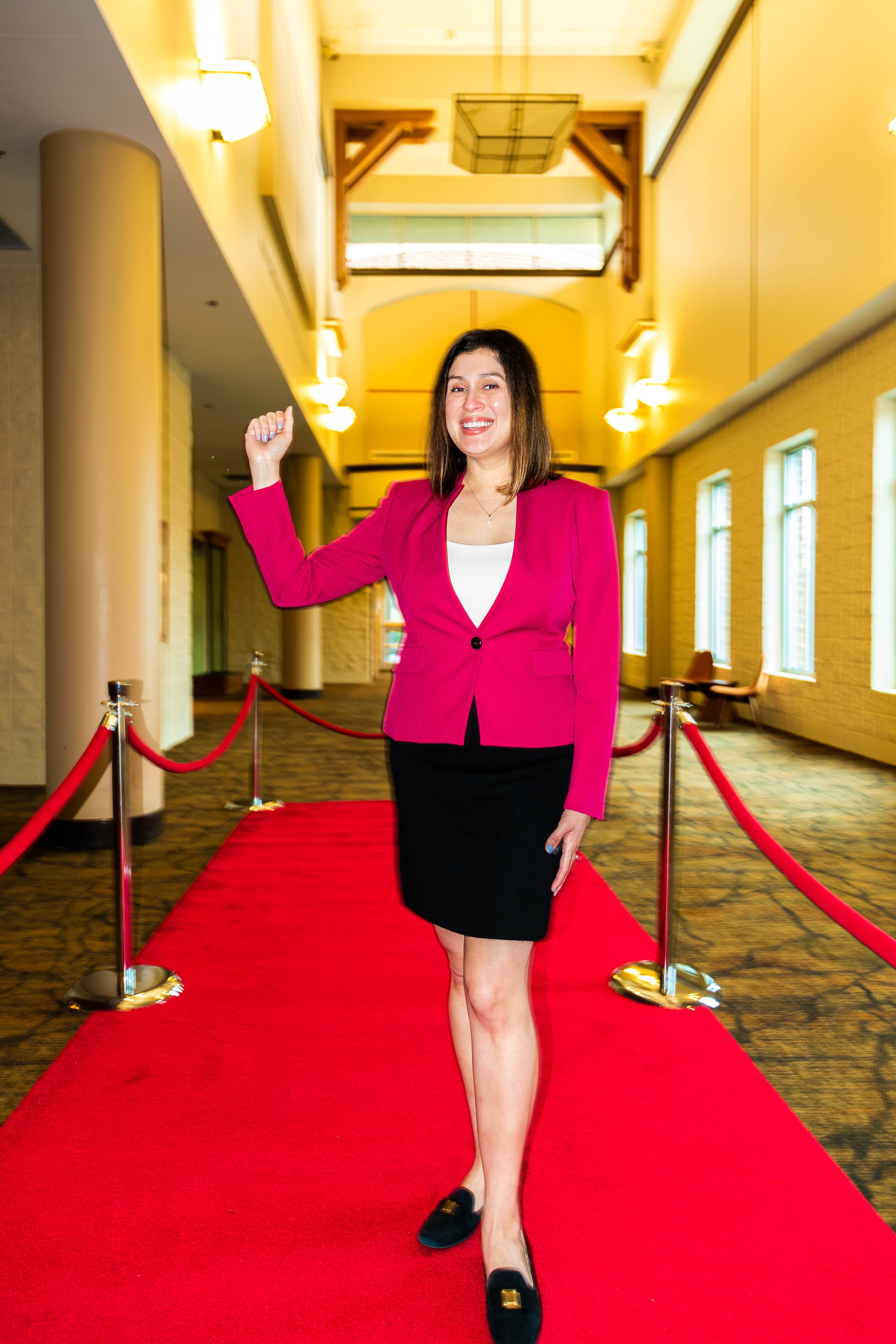 Yevette Lynn, brand photographer, captures an event-style portrait of a woman in a red blazer walking a red carpet inside a bright venue.