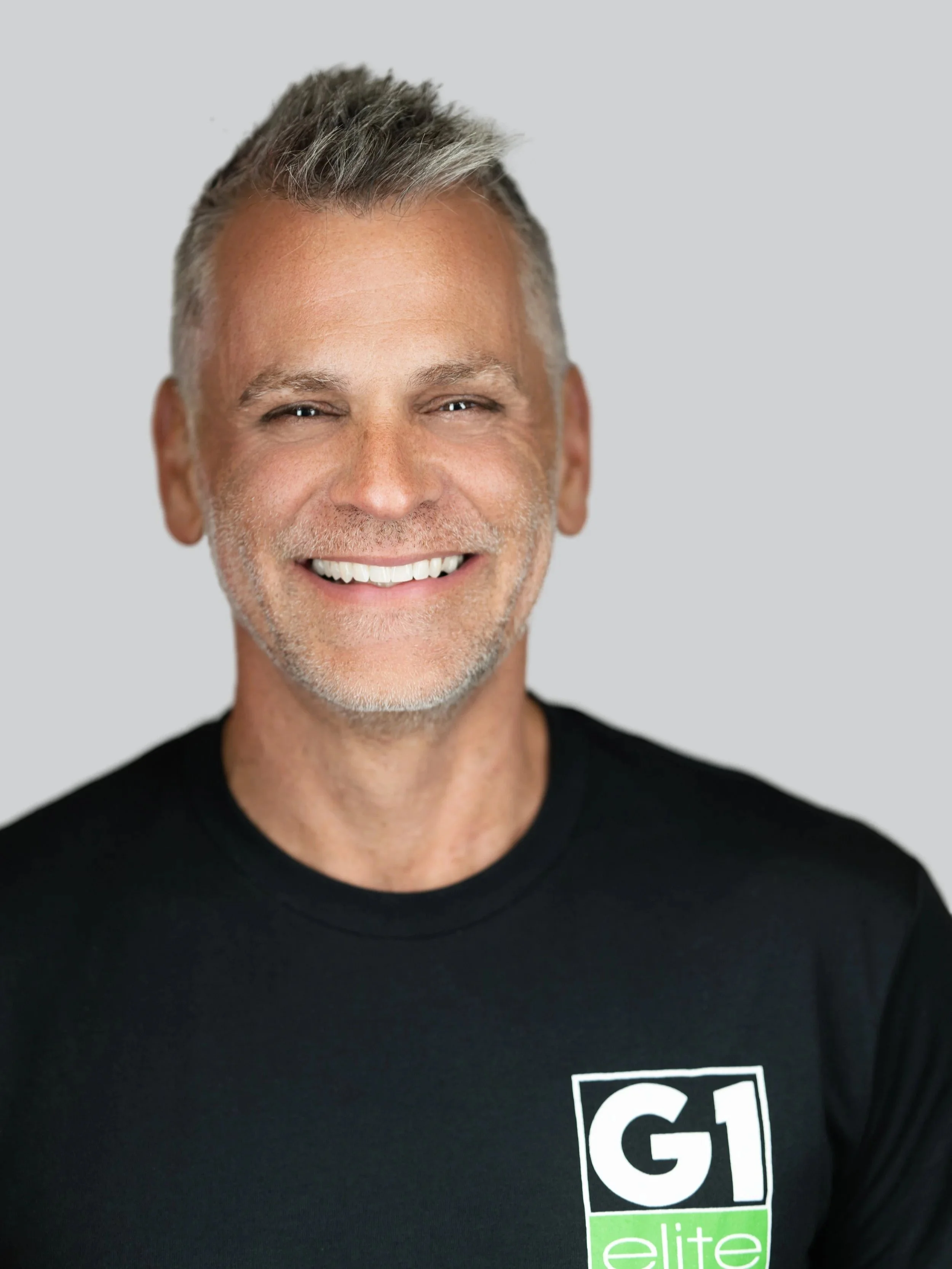 Professional headshot of a smiling male small business owner wearing a black G1 Elite company shirt against a light gray background.
