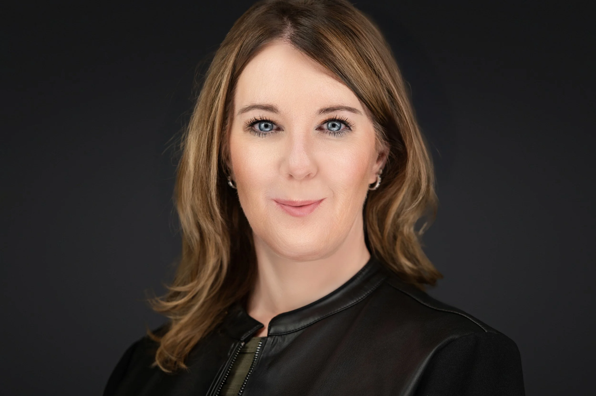 Yevette Lynn, professional photographer, creates a polished studio headshot of a woman wearing a black jacket with a soft expression against a dark background.