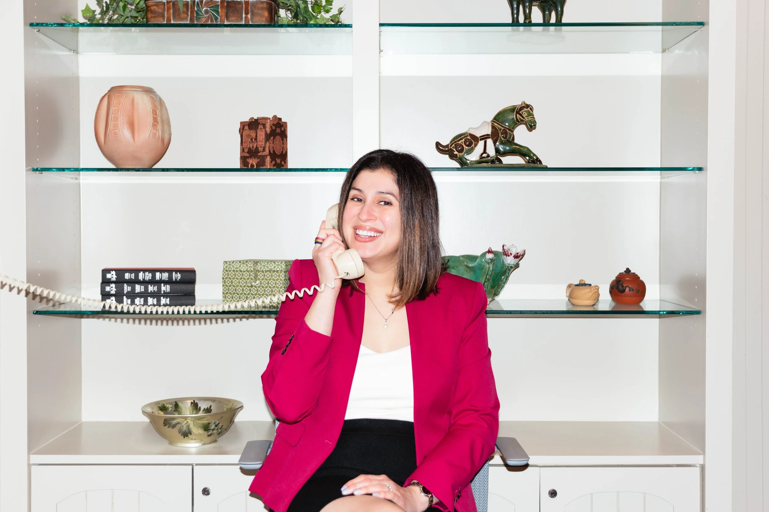 Yevette Lynn, brand photographer, captures a lifestyle office portrait of a woman in a red blazer smiling while holding a vintage desk phone.
