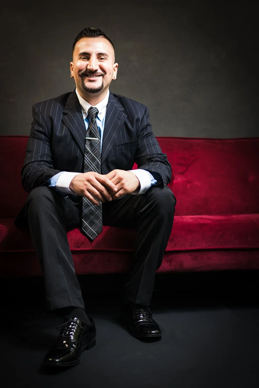 Yevette Lynn portrait photographer captures professional studio portrait of smiling man in suit, seated on red sofa against dark backdrop.