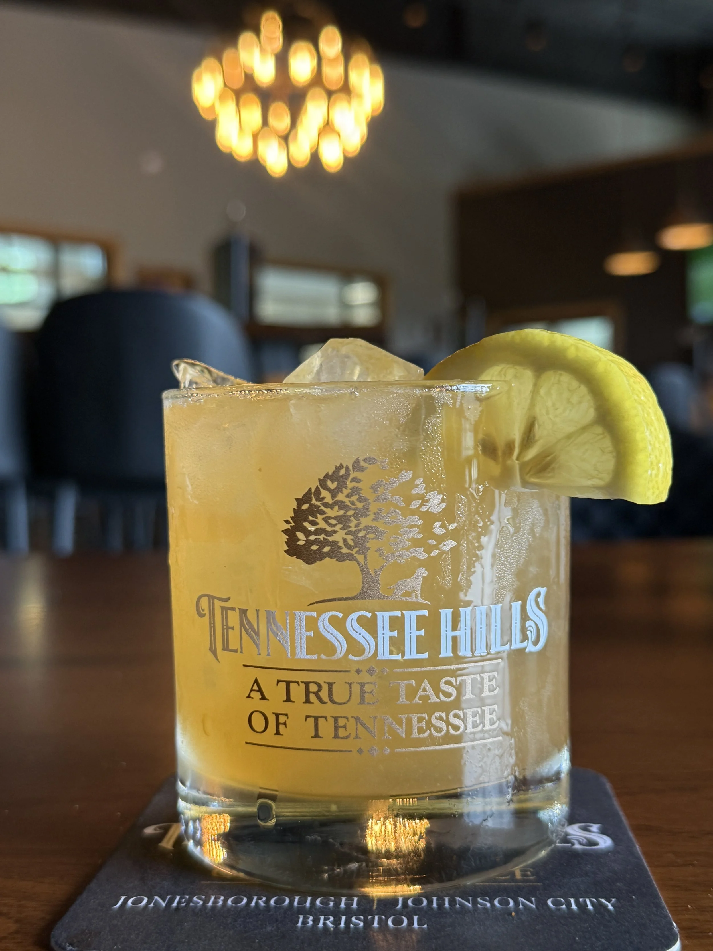 Cocktail with ice and lemon garnish in a Tennessee Hills glass on a bar table, with warm lights blurred in the background.