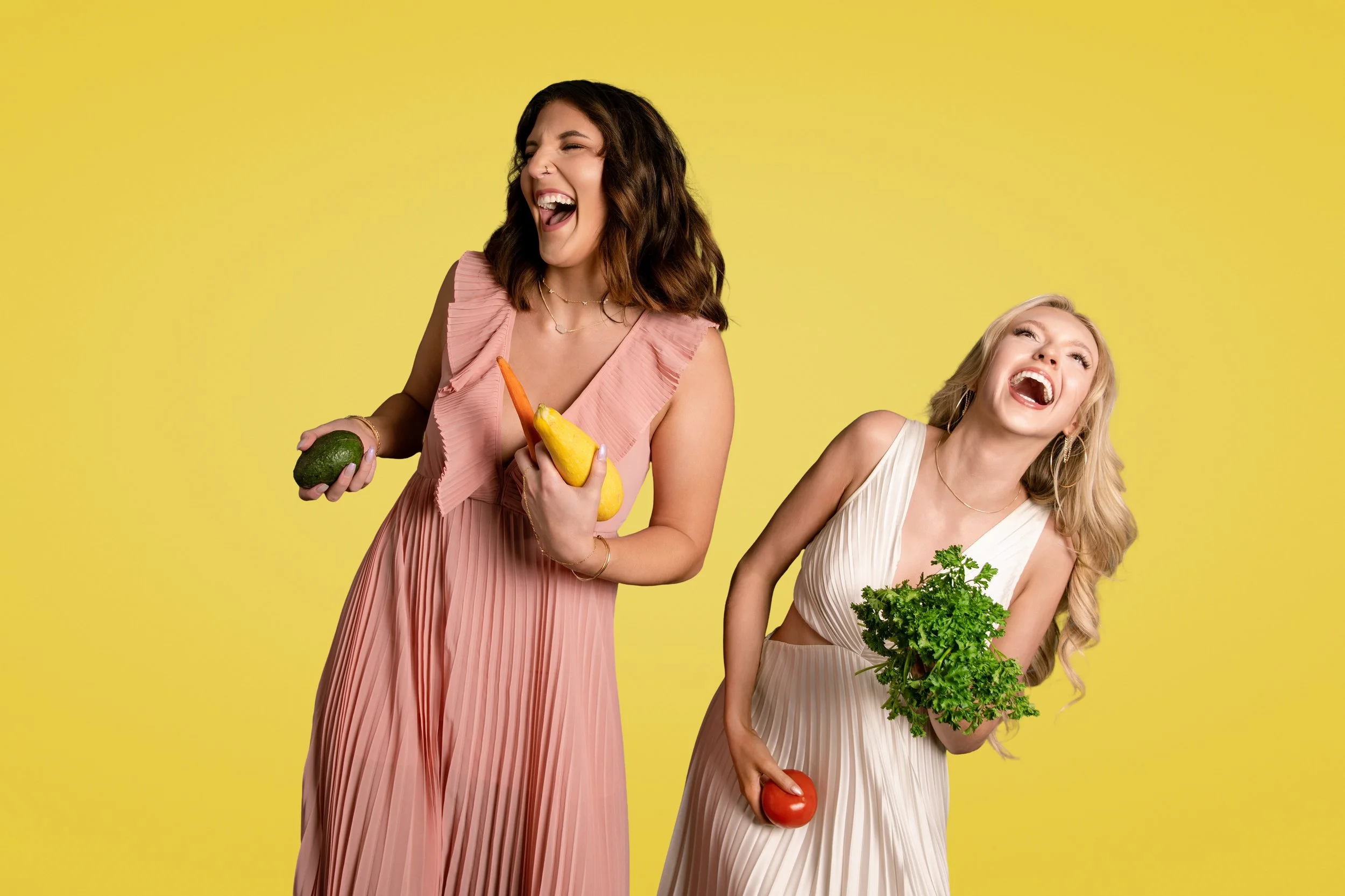 Yevette Lynn portrait photographer captures joyful studio lifestyle portrait of two women laughing while holding fresh vegetables against bright yellow background.