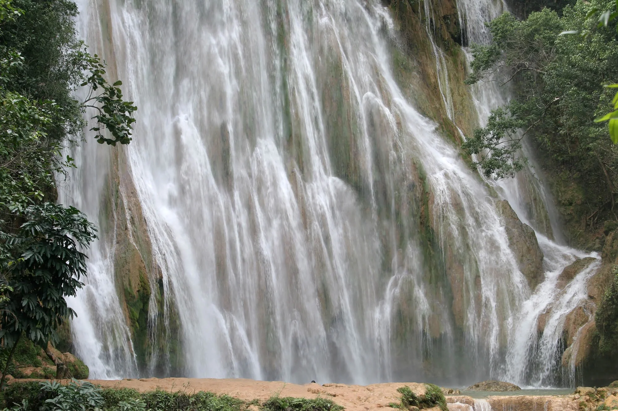 Large waterfall flowing over rocks surrounded by lush green trees and vegetation