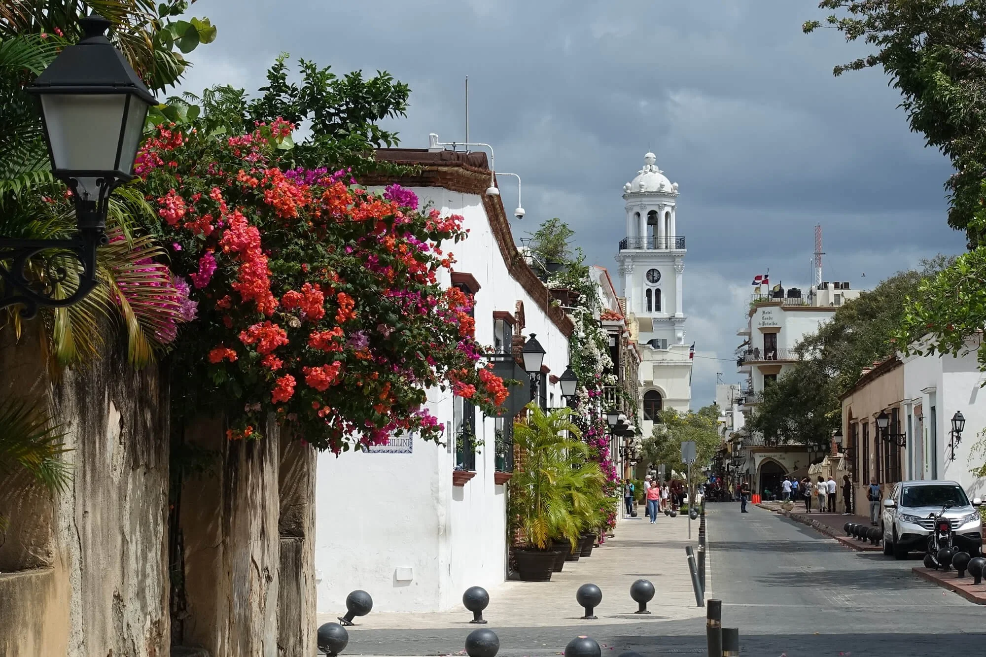A street scene with white buildings, colorful flowers, and a tall white clock tower in the background. There are people walking and cars parked along the side. Street lamps and potted plants line the sidewalk, and the sky is cloudy.