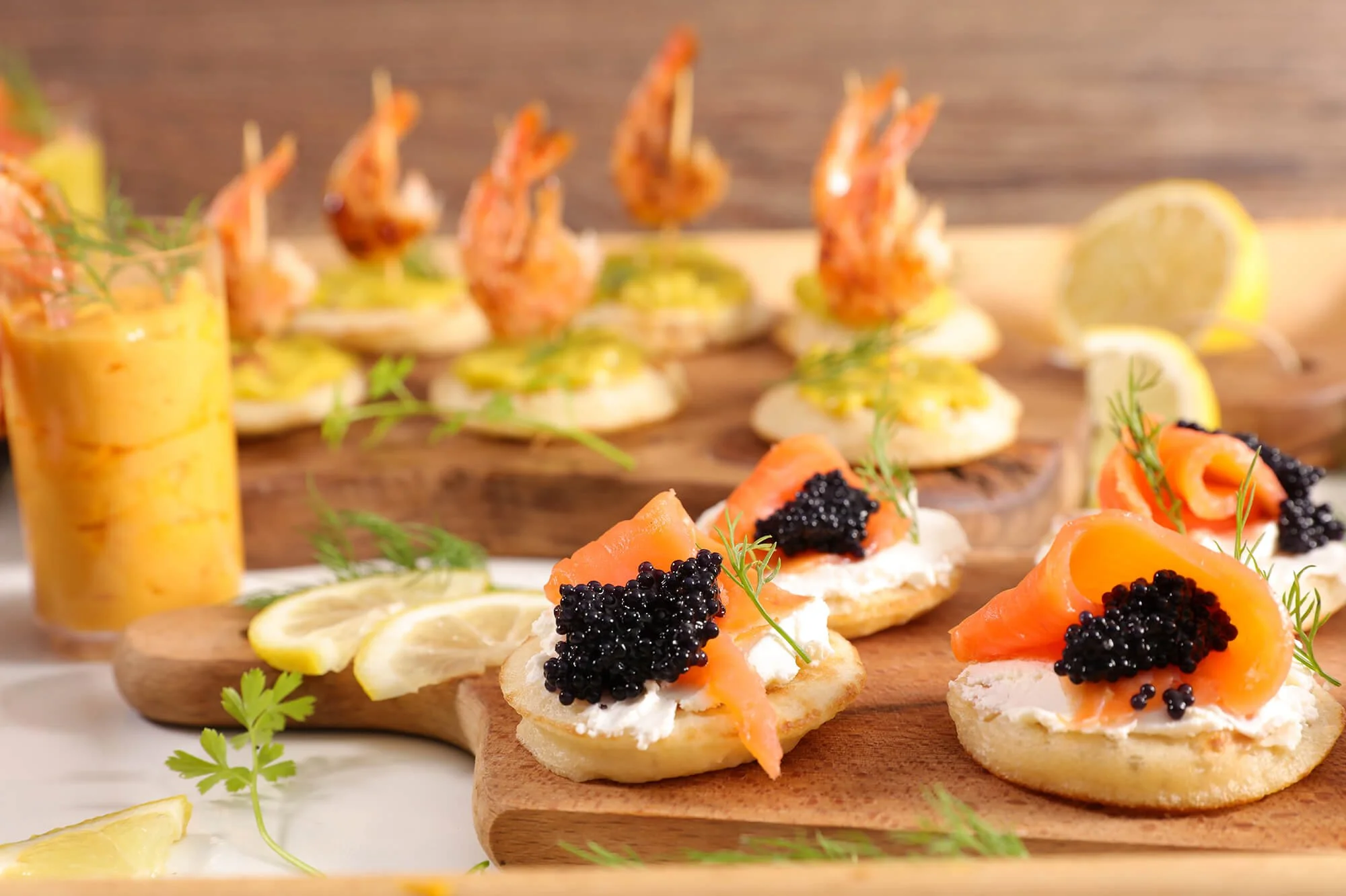 Close-up of smoked salmon and caviar canapés on a wooden platter, garnished with fresh herbs and lemon slices, with shrimp appetizers in the background.