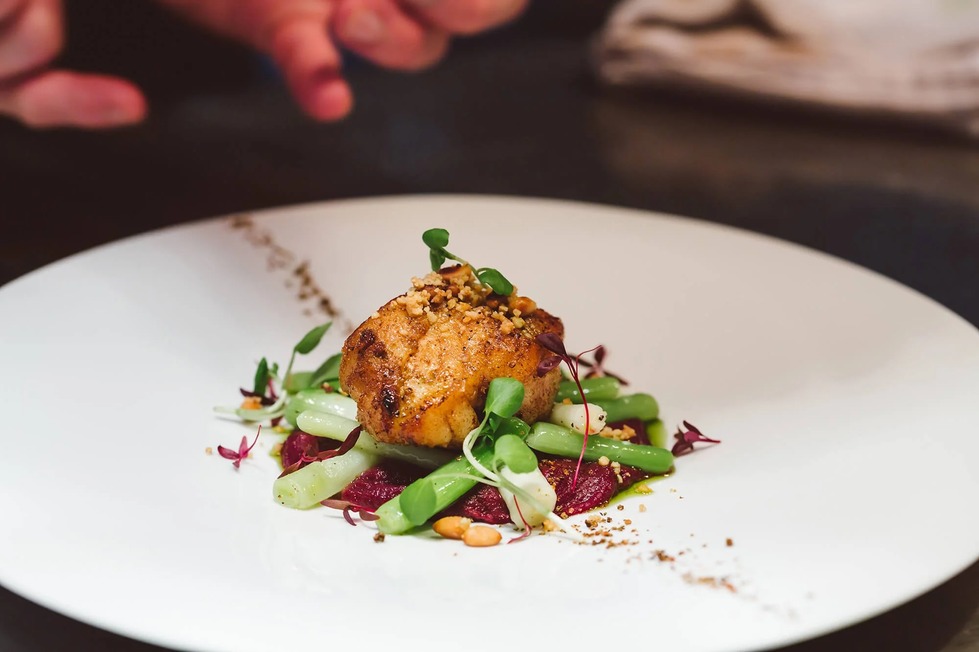 A plated gourmet dish featuring a seared piece of chicken on a bed of green beans, beets, microgreens, and garnished with crushed nuts and a sprinkle of ground spices.